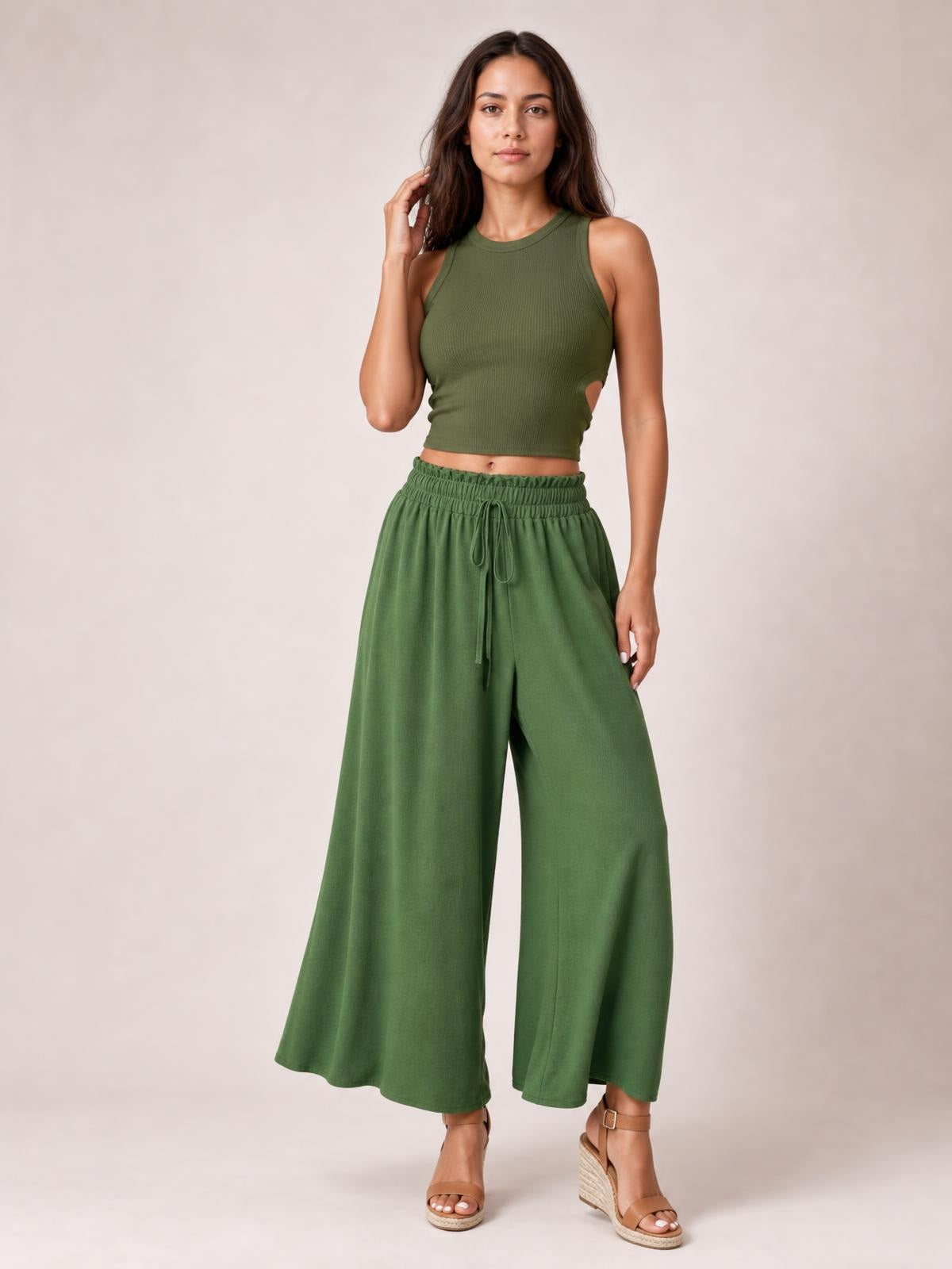 Model wearing olive green tied high waist wide leg pants with an elastic waistband and flowy, full-length silhouette, standing with one hand in her hair.