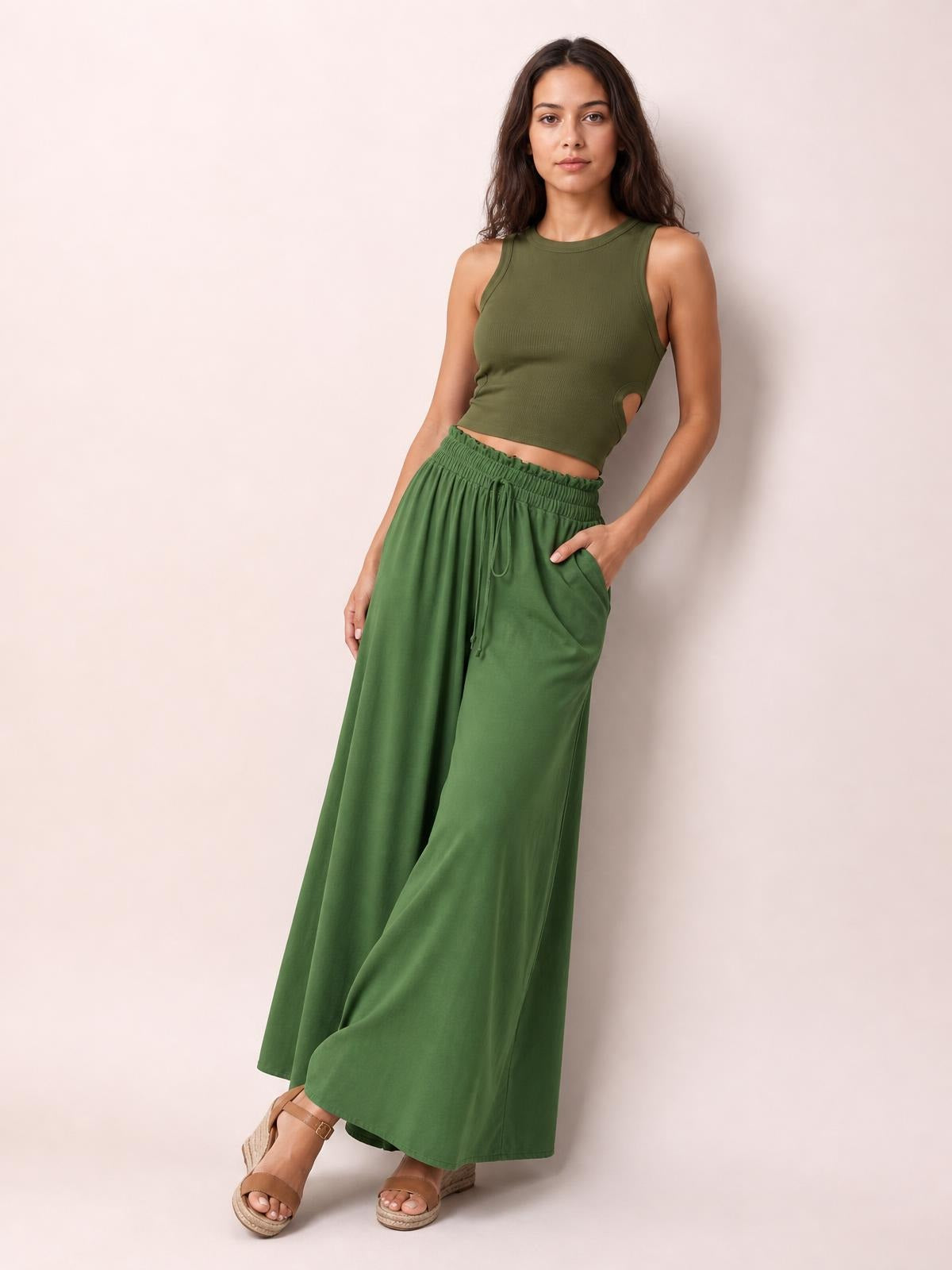 Model poses in green tied high waist wide leg pants with a gathered waistband, drawstring, side pockets, and a flowing full-length silhouette.