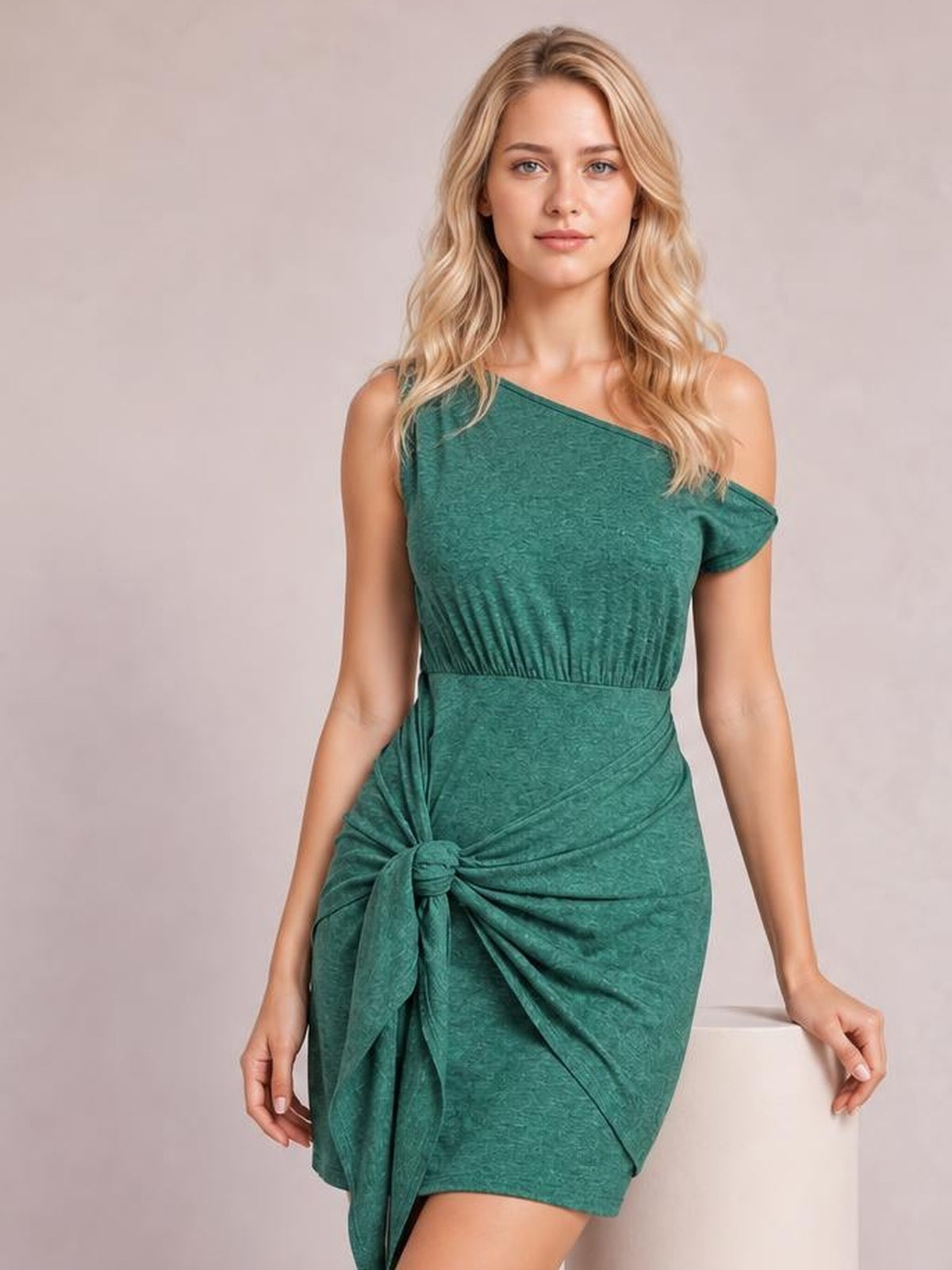 A model poses in a fitted green single-shoulder sleeveless mini dress with a ruched bodice, wide waistband, and tied hip detail.