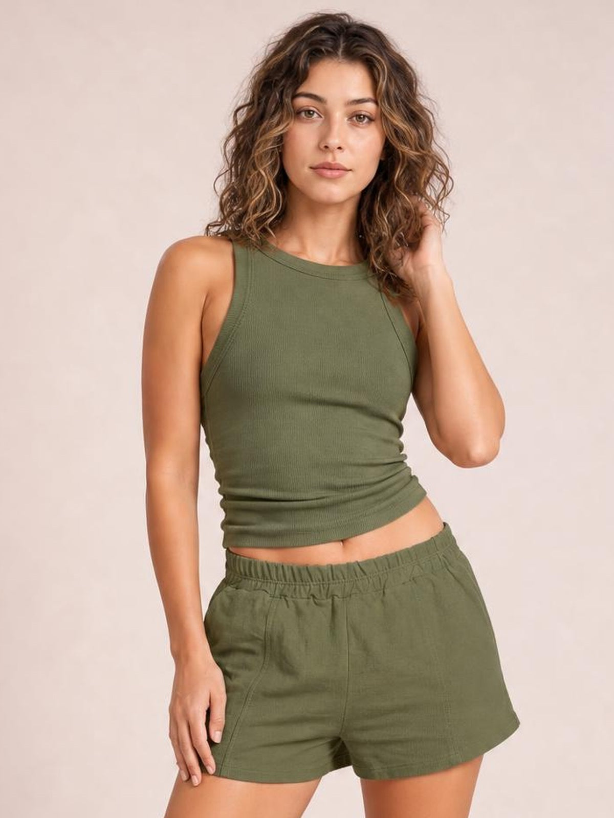 A woman modeling an olive green two-piece lounge set consisting of a sleeveless crop top and matching drawstring shorts, set against a solid light beige background.