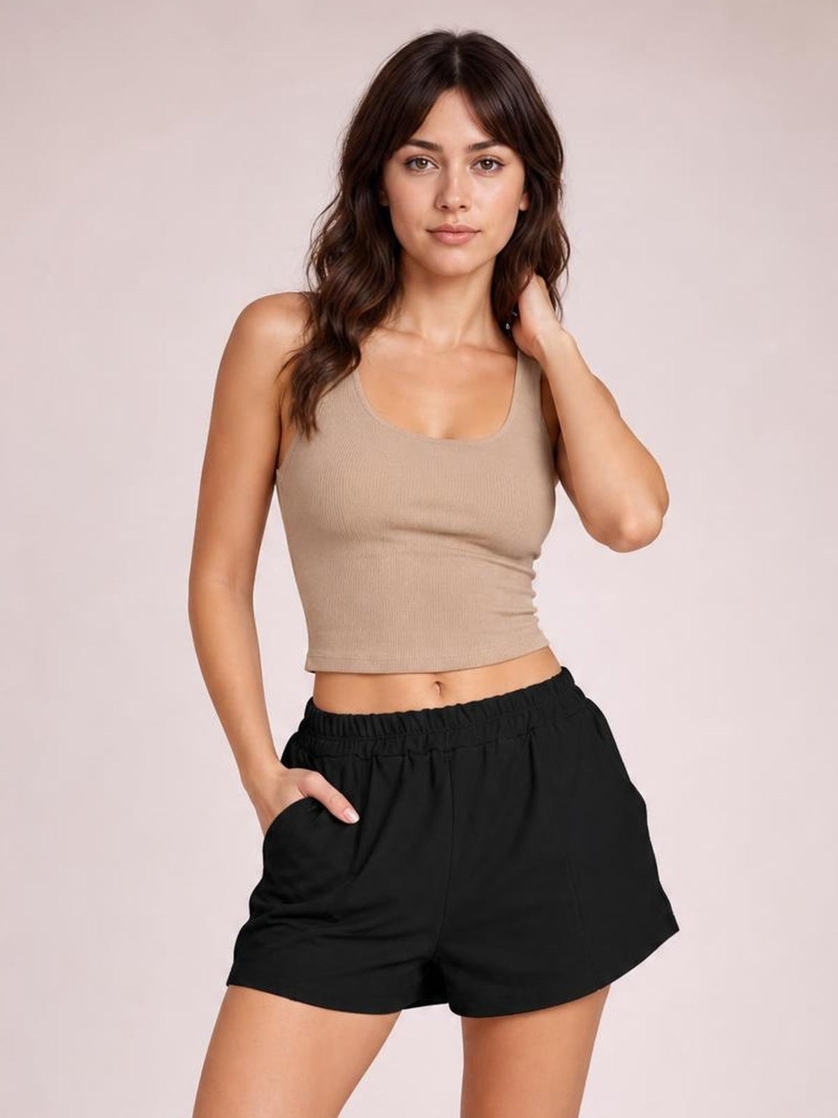 A woman modeling a beige ribbed crop tank top and black elastic-waist shorts against a solid light beige studio background.