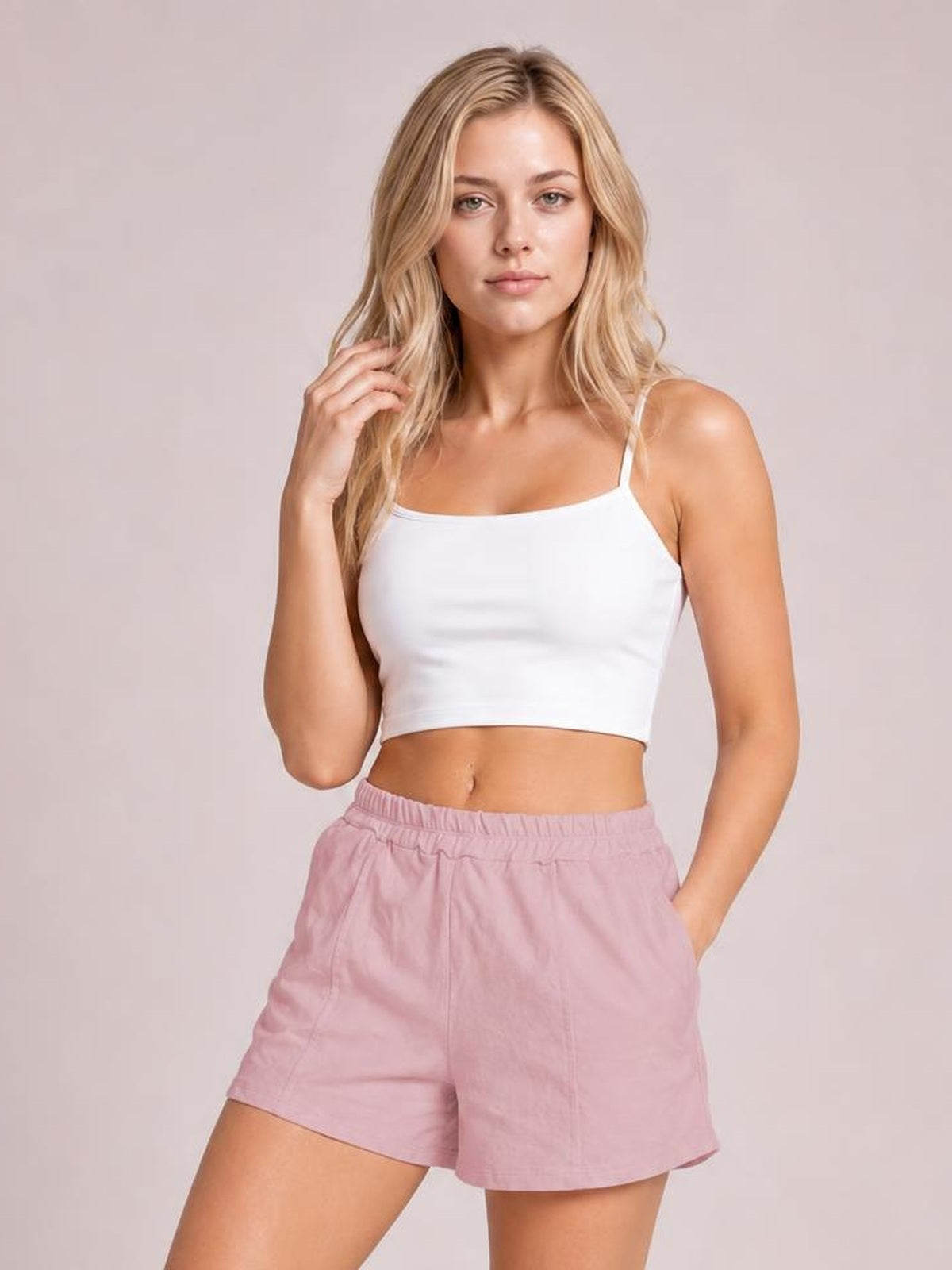 A woman modeling a white ribbed spaghetti-strap crop top and dusty pink shorts against a solid light beige studio background.