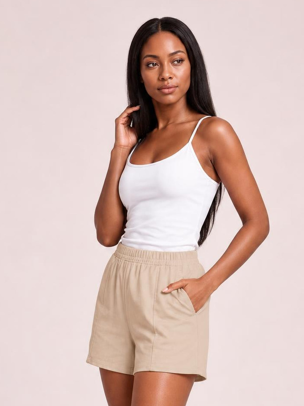 A woman modeling a white spaghetti-strap tank top and beige drawstring shorts against a solid pale pink studio background.