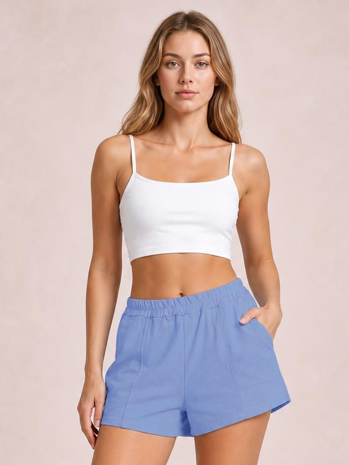 A woman modeling a white spaghetti-strap crop top and light blue shorts against a soft beige studio background.