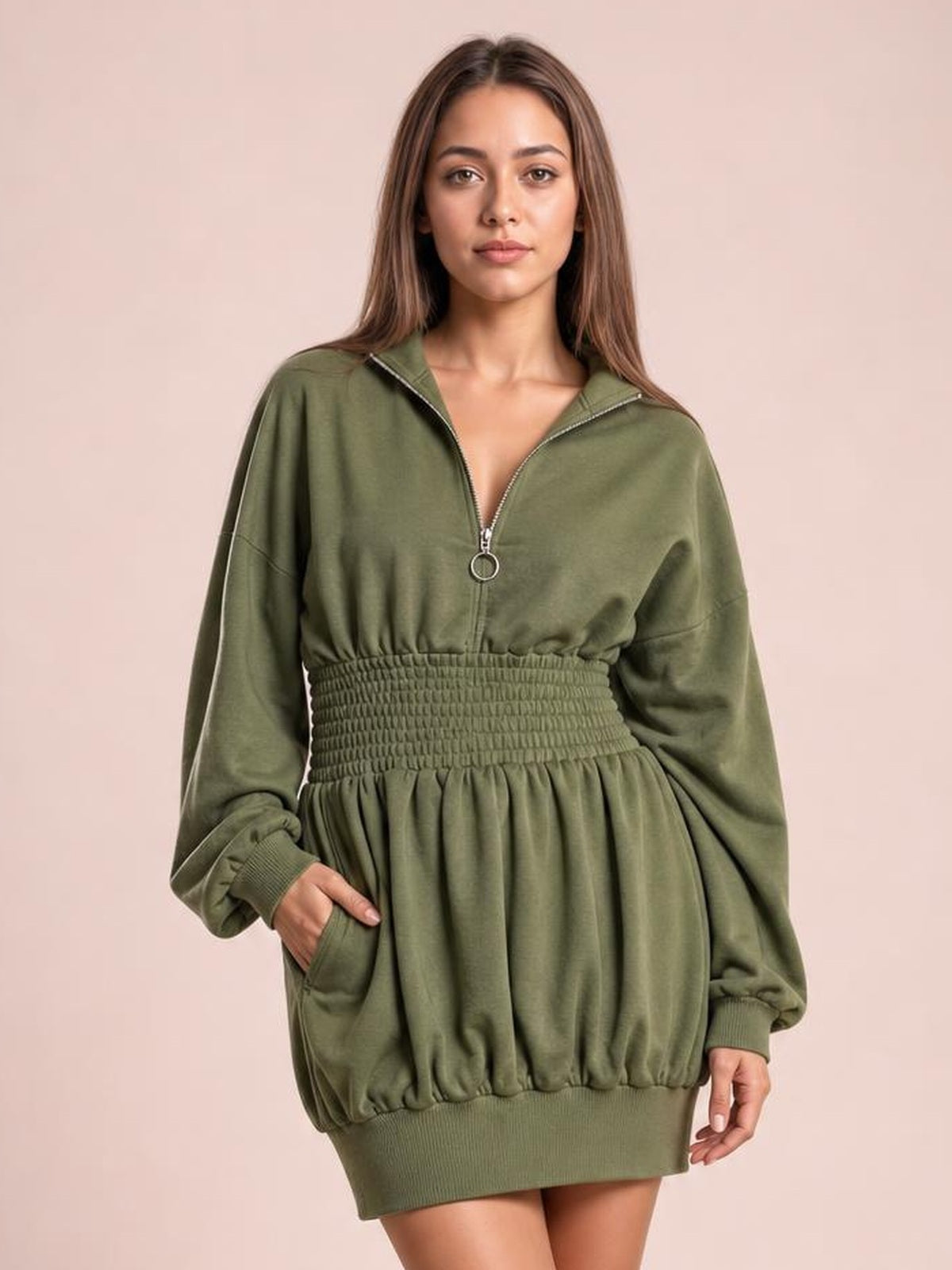 A model poses in an olive green hooded long sleeve mini dress with a smocked waist, front zipper, relaxed silhouette, ribbed cuffs and hem, and side pockets.
