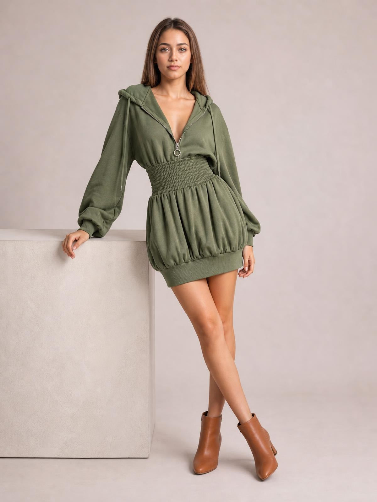 Model poses in a sage green hooded long sleeve mini dress with a front zipper, drawstring, smocked waist, and gathered bubble skirt.