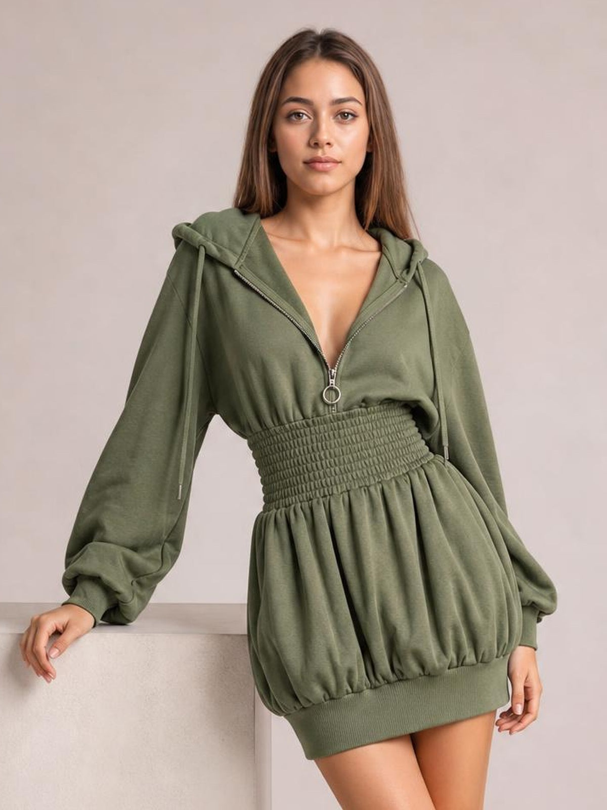 A model leans against a ledge wearing an olive green hooded long sleeve mini dress with a zip front, drawstrings, smocked waist, and relaxed silhouette.