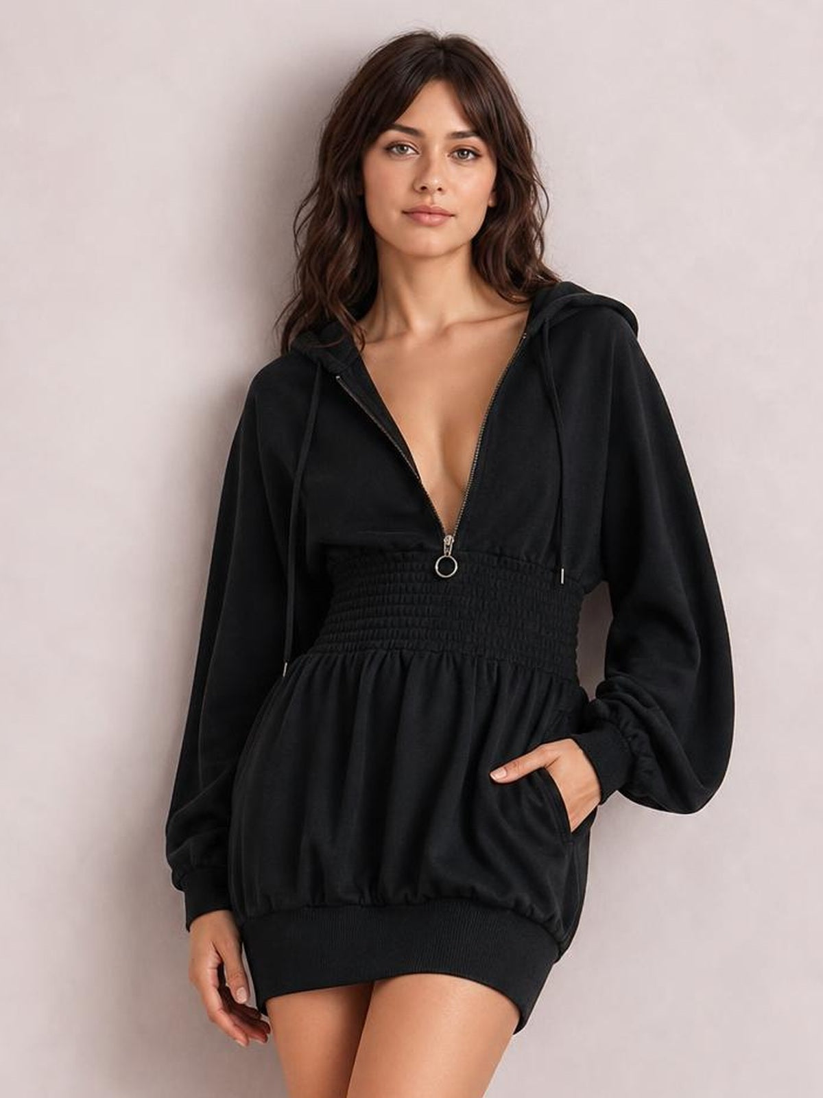 A model stands in a black hooded long-sleeve mini dress with a front zipper, drawstrings, smocked waist, relaxed silhouette, and visible side pocket.