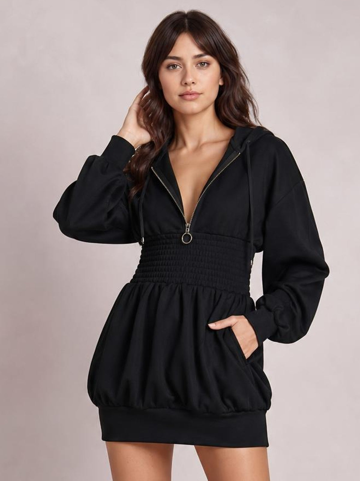 A model poses in a black hooded long-sleeve mini dress with a front zipper, smocked waist, gathered skirt, ribbed hem, and pockets.
