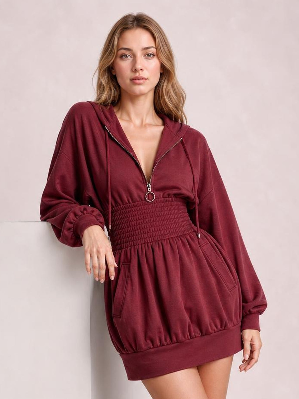 Model leans against a wall wearing a burgundy hooded long-sleeve mini dress with a zip front, drawstrings, smocked waist, and side pockets.