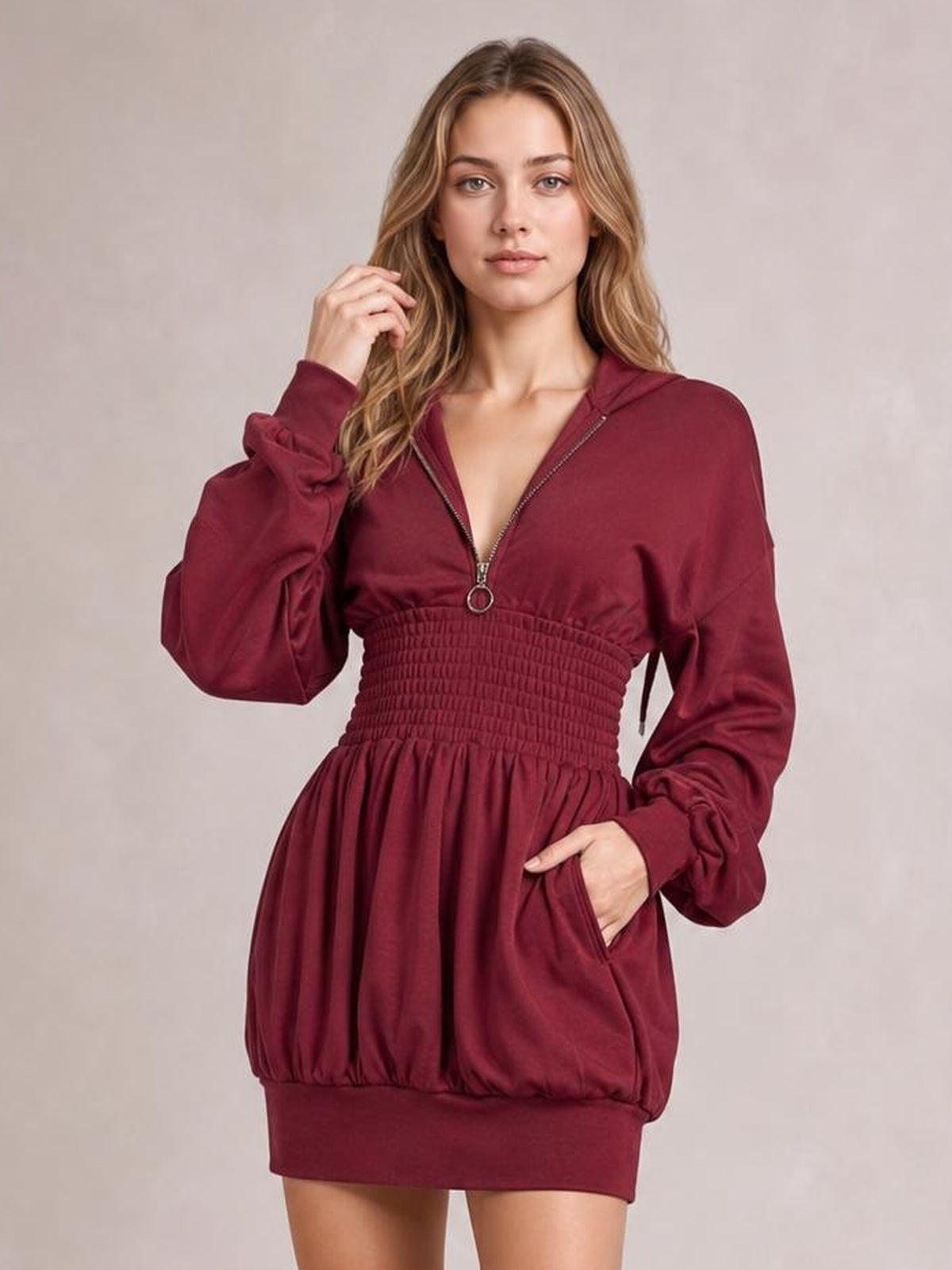A model poses in a burgundy hooded long-sleeve mini dress with a front zipper, smocked waist, gathered skirt, ribbed hem, and side pockets.