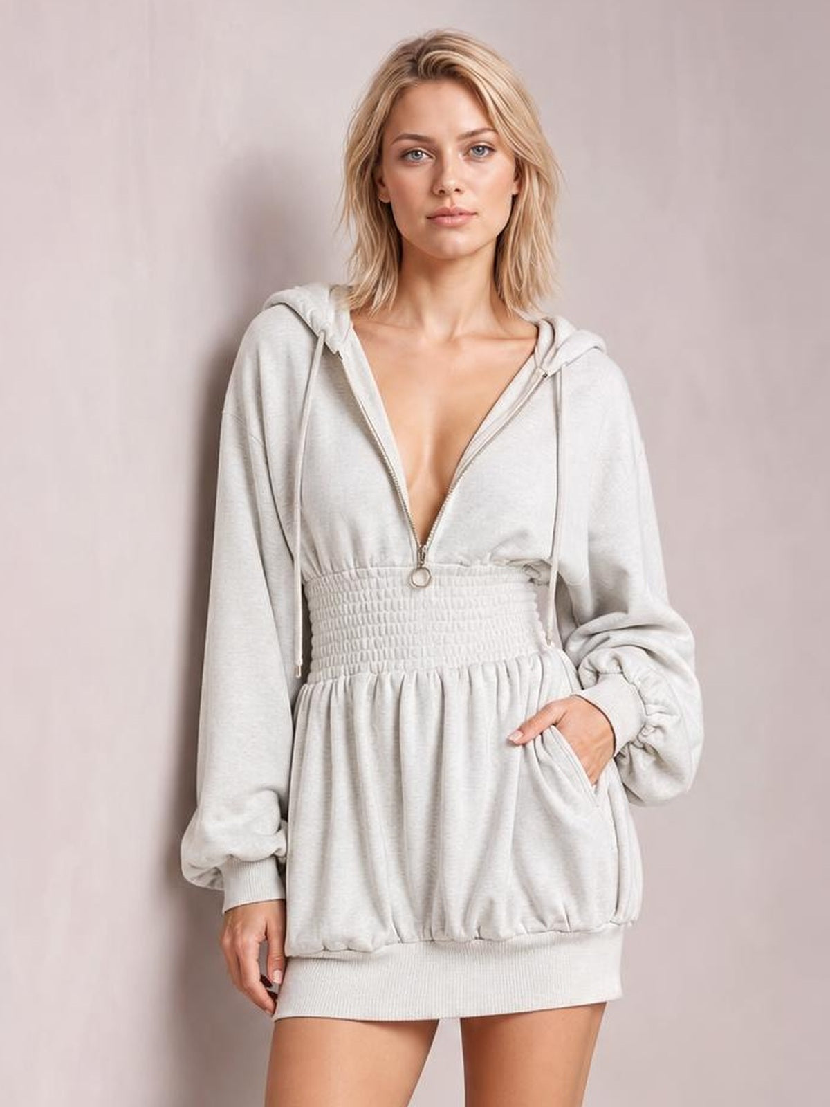 A model wears a light gray hooded long sleeve mini dress with a front zipper, drawstrings, smocked waist, relaxed silhouette, and side pocket.