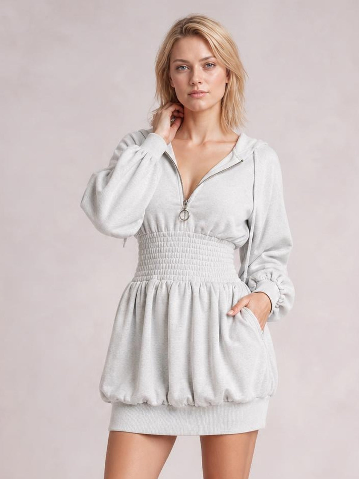 A model poses in a light gray hooded long-sleeve mini dress with a half-zip front, smocked waist, gathered skirt, and side pockets.
