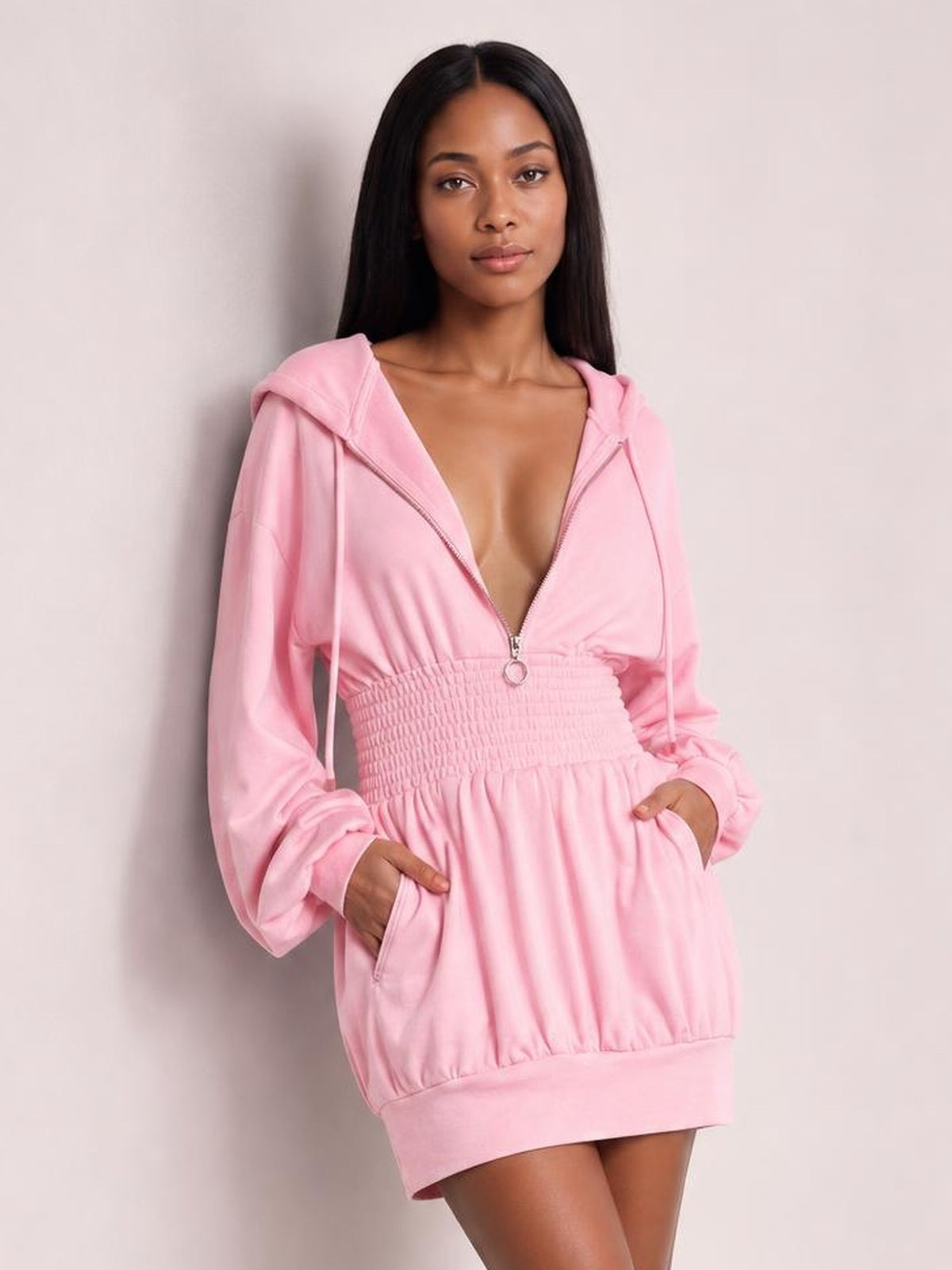 A model wears a pink hooded long-sleeve mini dress with a front zipper, drawstrings, smocked waist, relaxed silhouette, and side pockets.