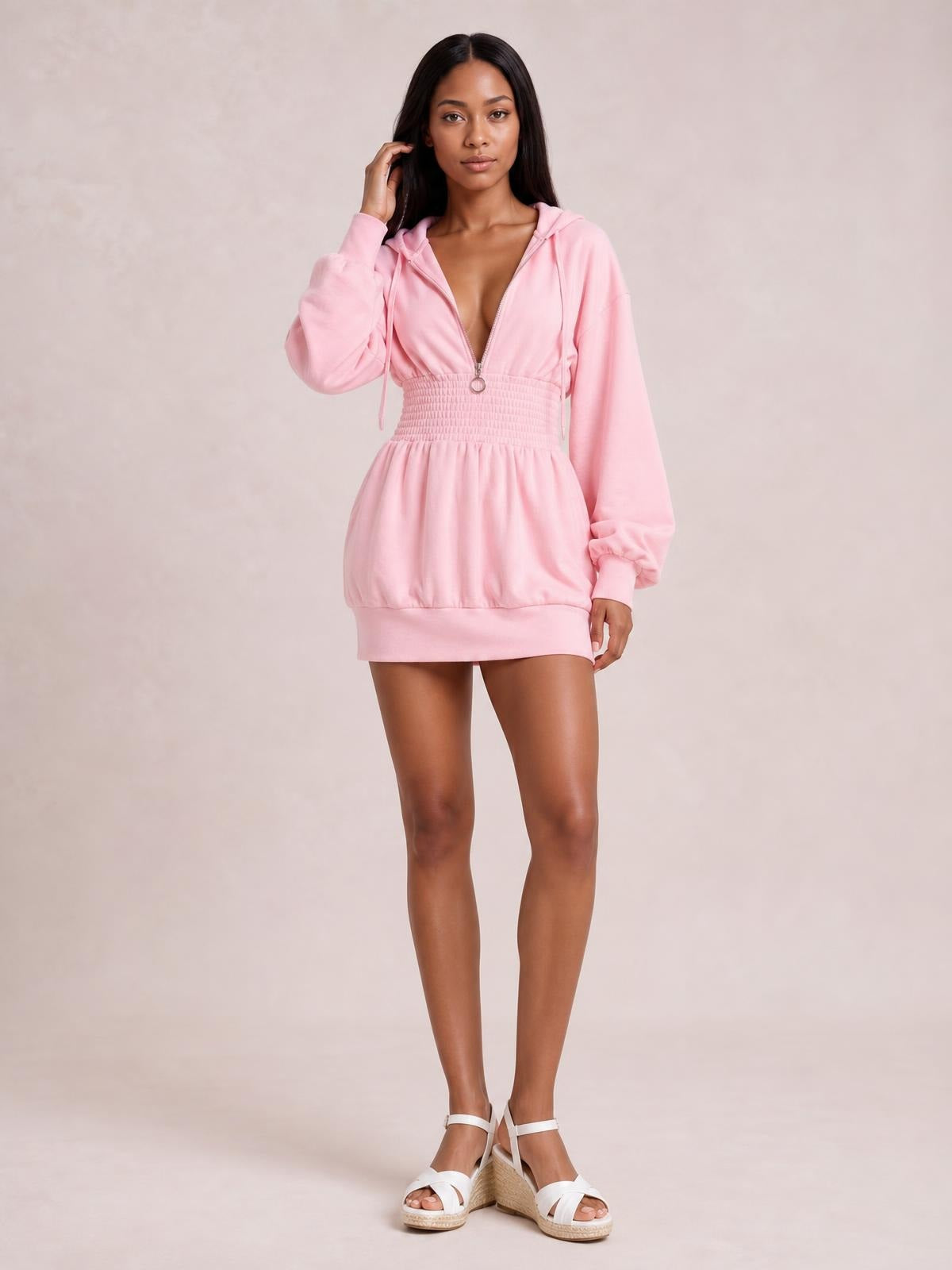 A model poses in a pink hooded long sleeve mini dress with a front zipper, drawstring hood, smocked waist, and relaxed, gathered silhouette.