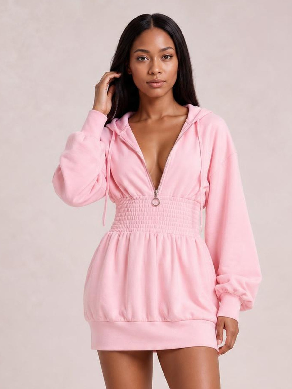 Model touches her hair while wearing a pink hooded long-sleeve mini dress with a zip front, drawstrings, smocked waist, and gathered skirt.