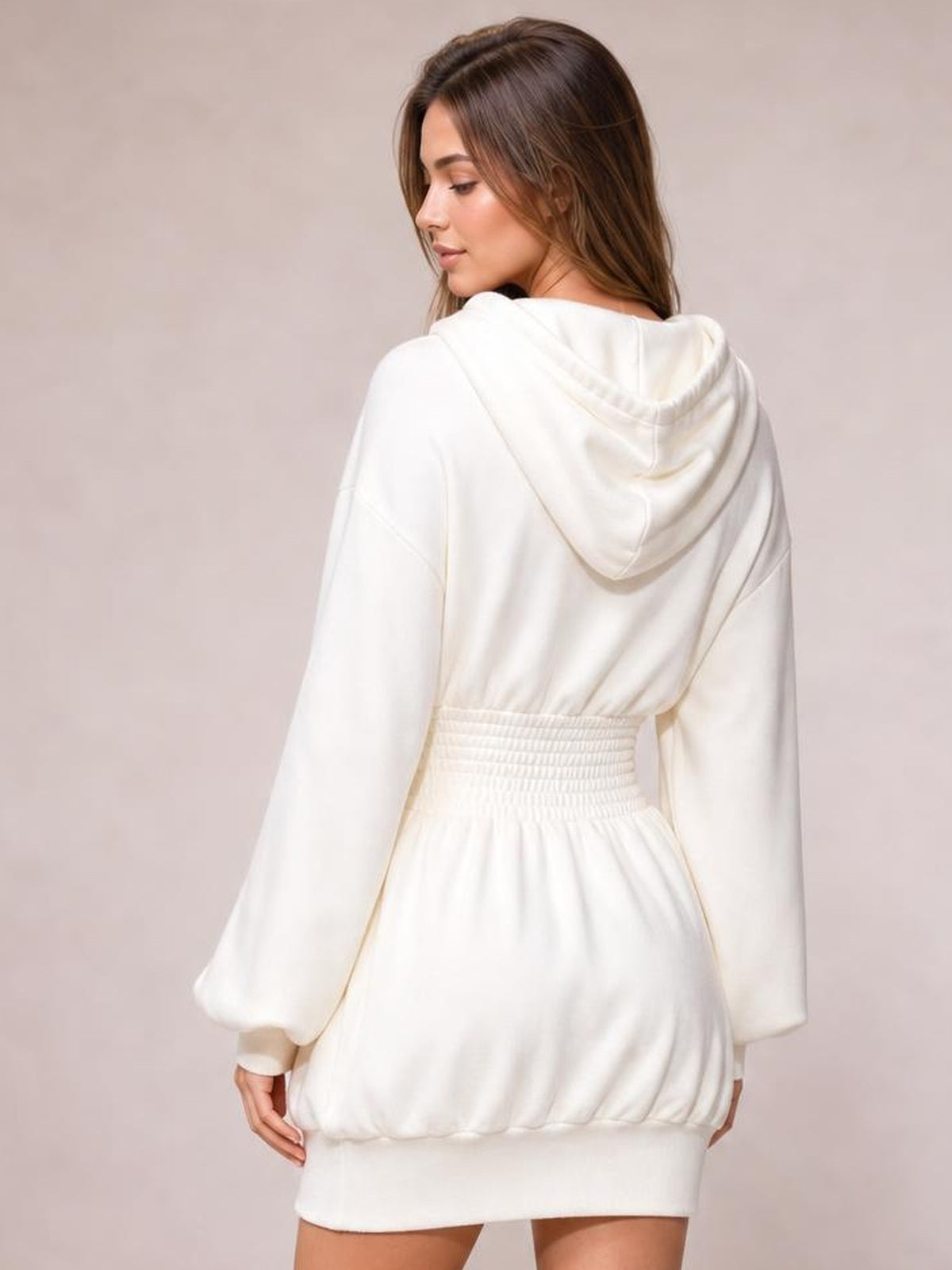 A model looks over her shoulder wearing a white hooded long-sleeve mini dress with a smocked waist, relaxed silhouette, and ribbed hem.