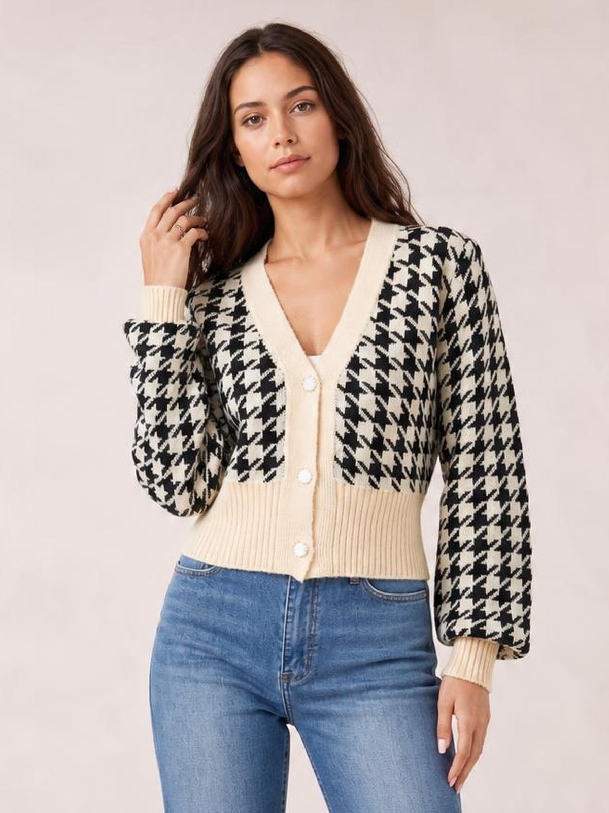 Model wearing a black-and-cream houndstooth button-up long sleeve cardigan with a V-neck, ribbed trim, fitted silhouette, and hand raised to her hair.