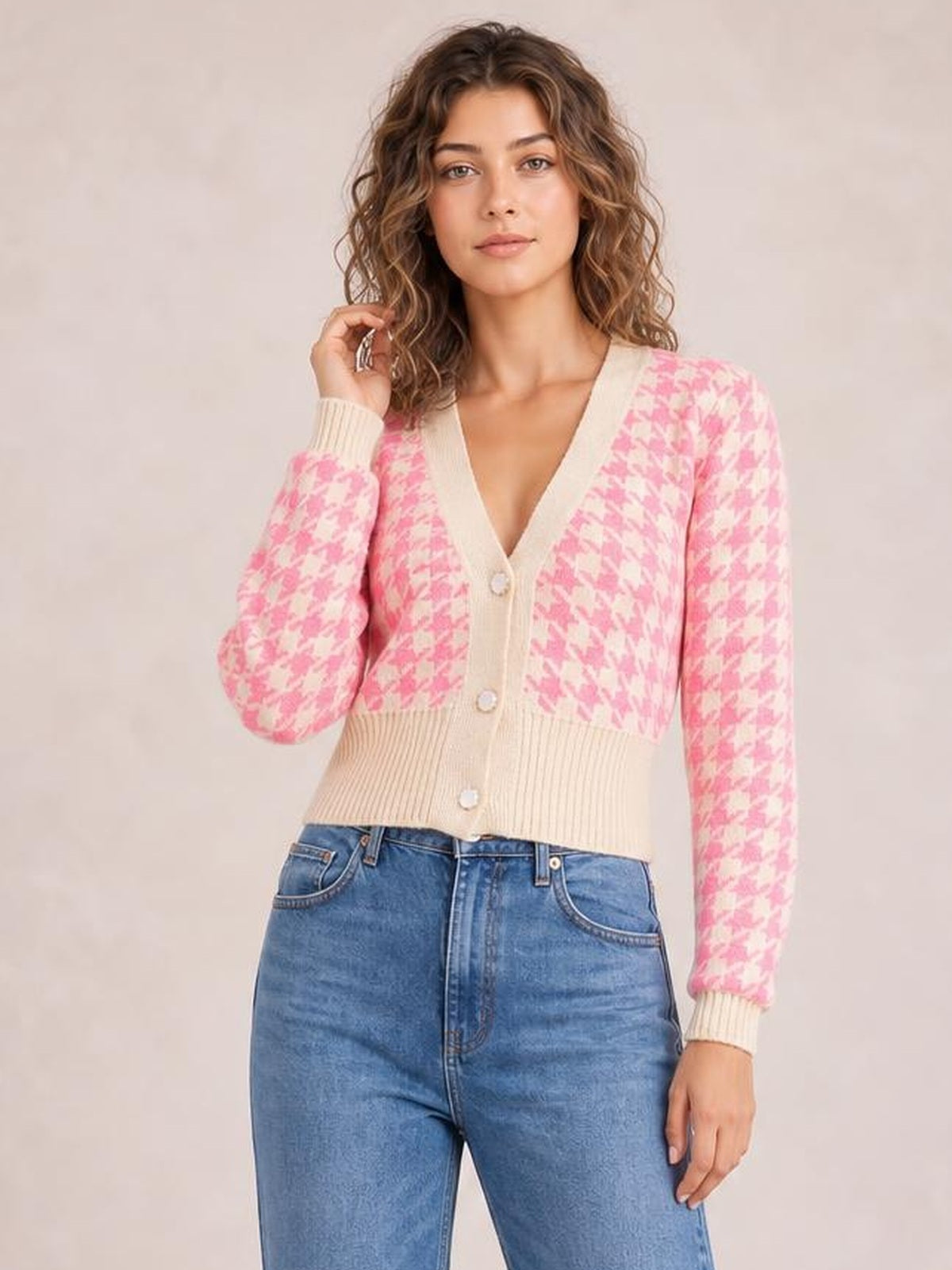 Model poses with one hand in her hair, wearing a pink and cream houndstooth button-up long-sleeve cardigan with a fitted silhouette and ribbed trim.