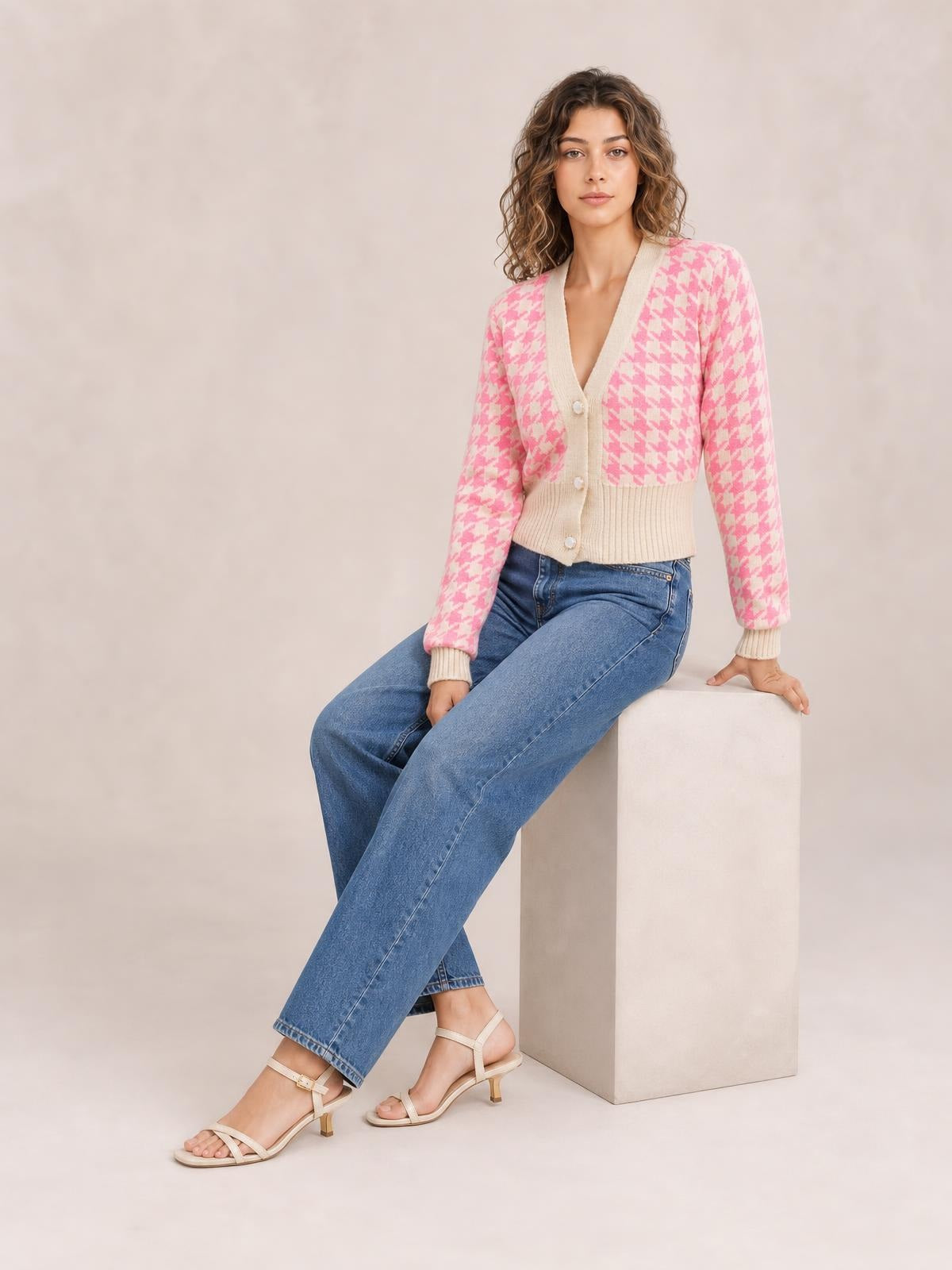 A seated model wears a pink and cream houndstooth button-up long-sleeve cardigan with a V-neck, ribbed trim, and fitted cropped silhouette.