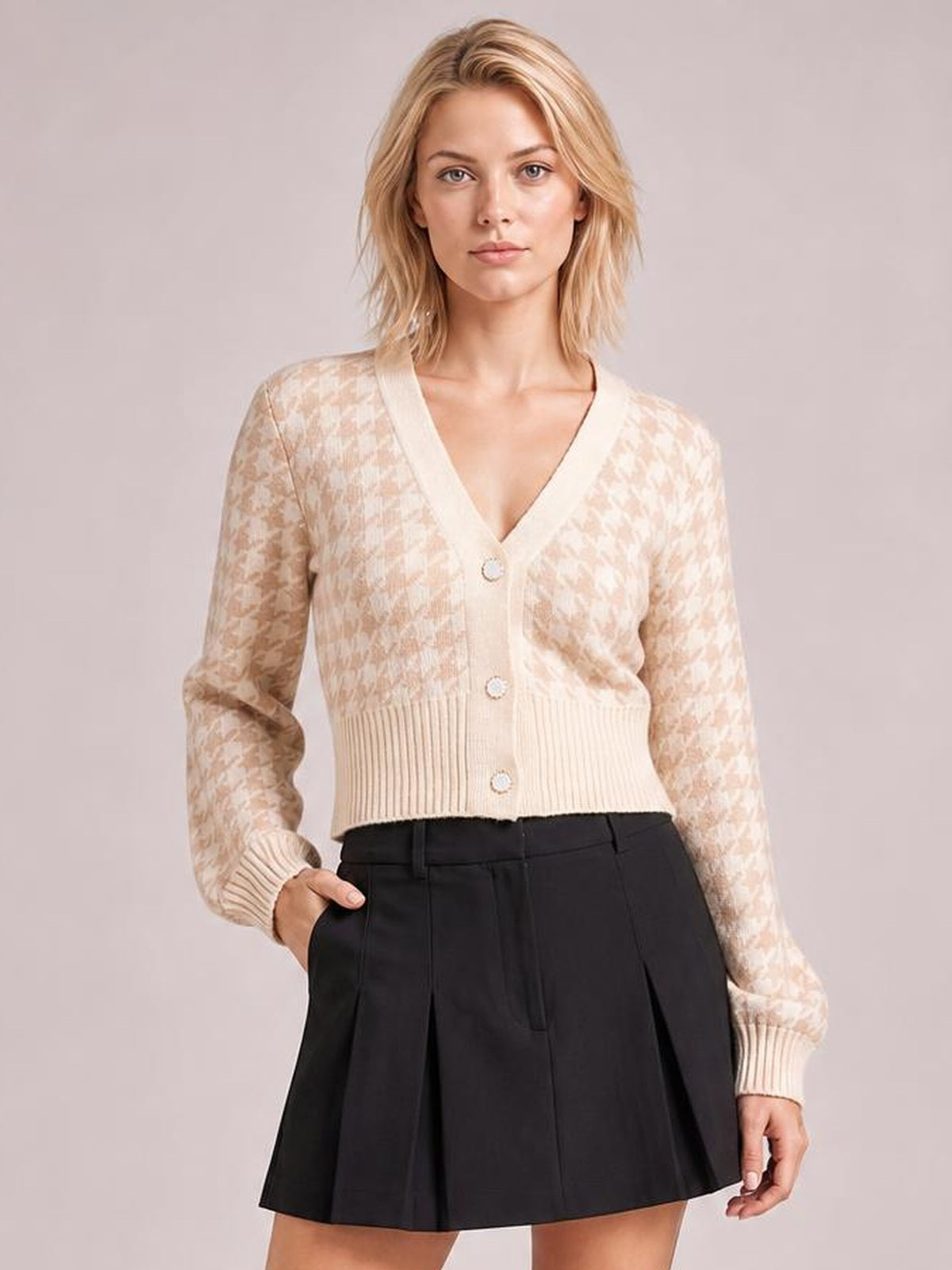 Model wearing a cream houndstooth button-up cardigan with long sleeves, V-neck, ribbed trim, and a fitted cropped silhouette, posing with one hand in her pocket.