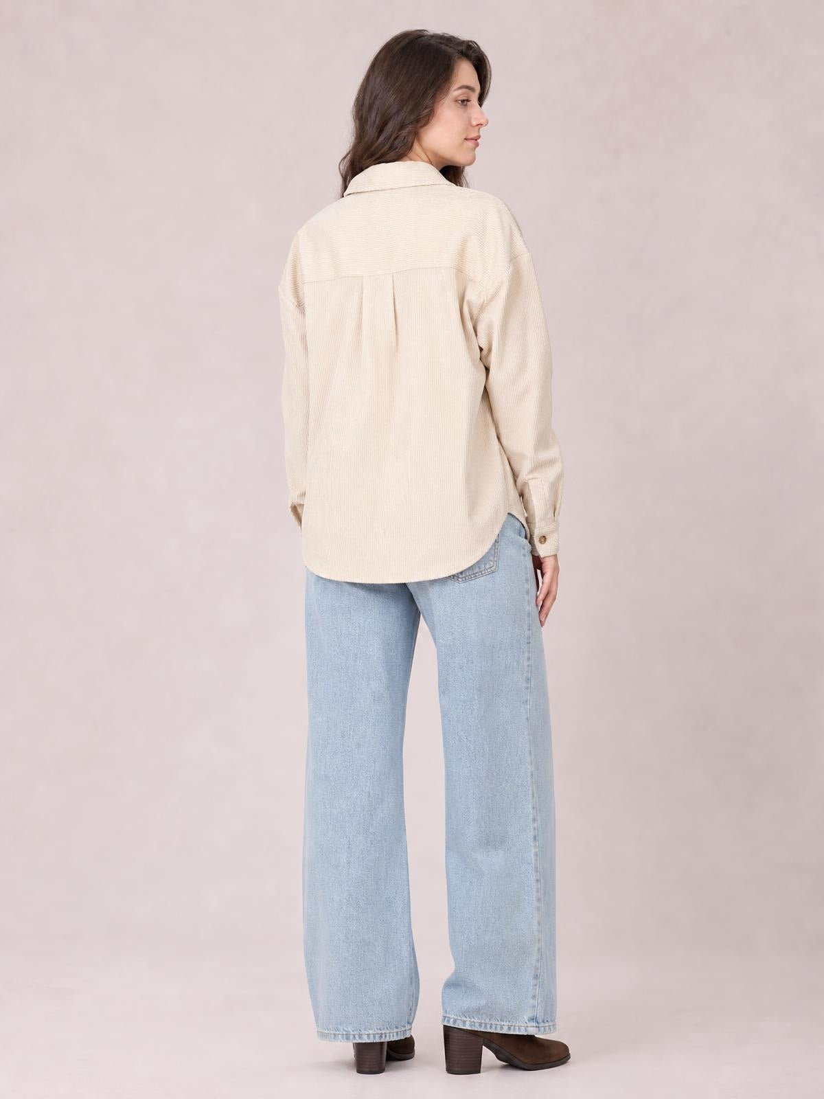 A model looks over her shoulder in a cream button-up long sleeve jacket with a relaxed silhouette, back yoke pleats, curved hem, and cuffed sleeves.