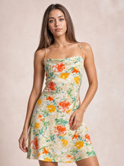 Model poses in a floral cowl neck satin mini dress with spaghetti straps, vibrant orange, yellow, coral, and green flowers, and a softly flared silhouette.