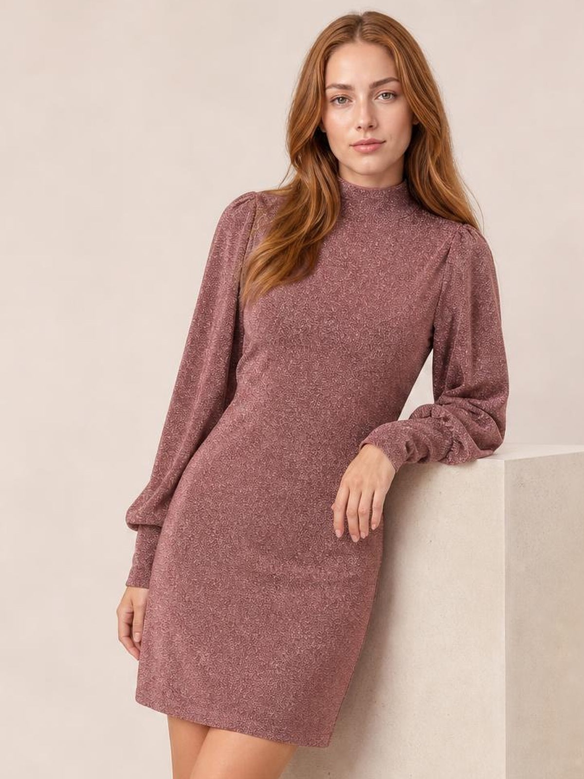 A model leans against a pedestal wearing a mauve glitter mock neck mini dress with long bishop sleeves and a figure-hugging silhouette.