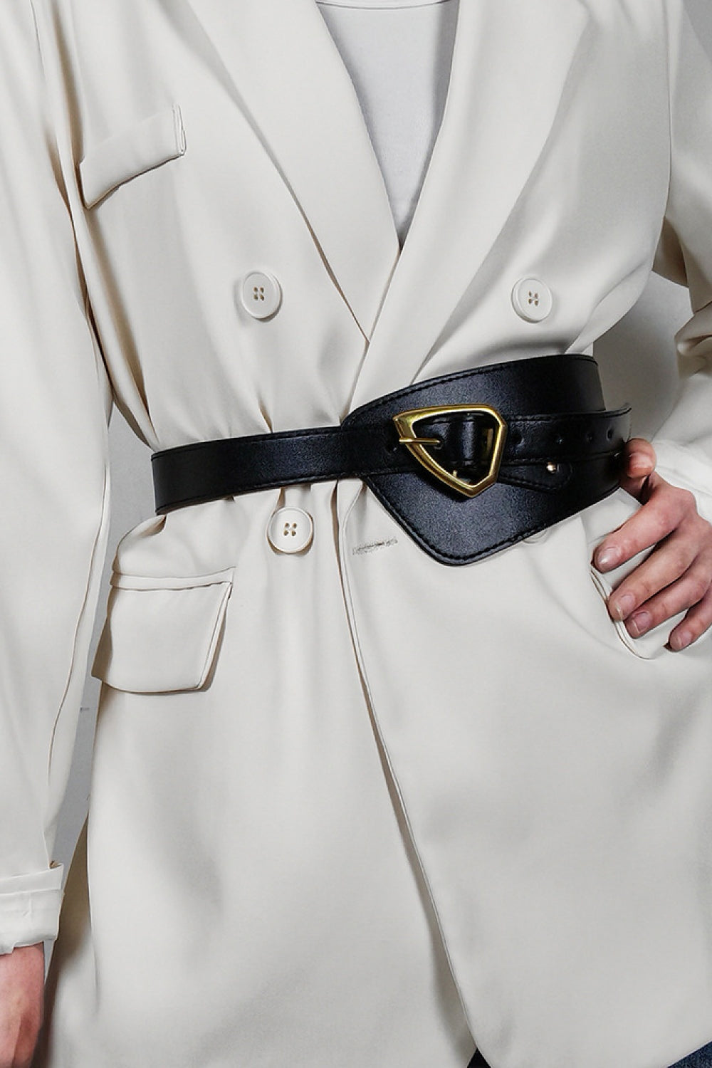 A model poses in a white blazer wearing a black irregular PU leather belt with a wide sculptural waist panel, narrow strap, and gold buckle.