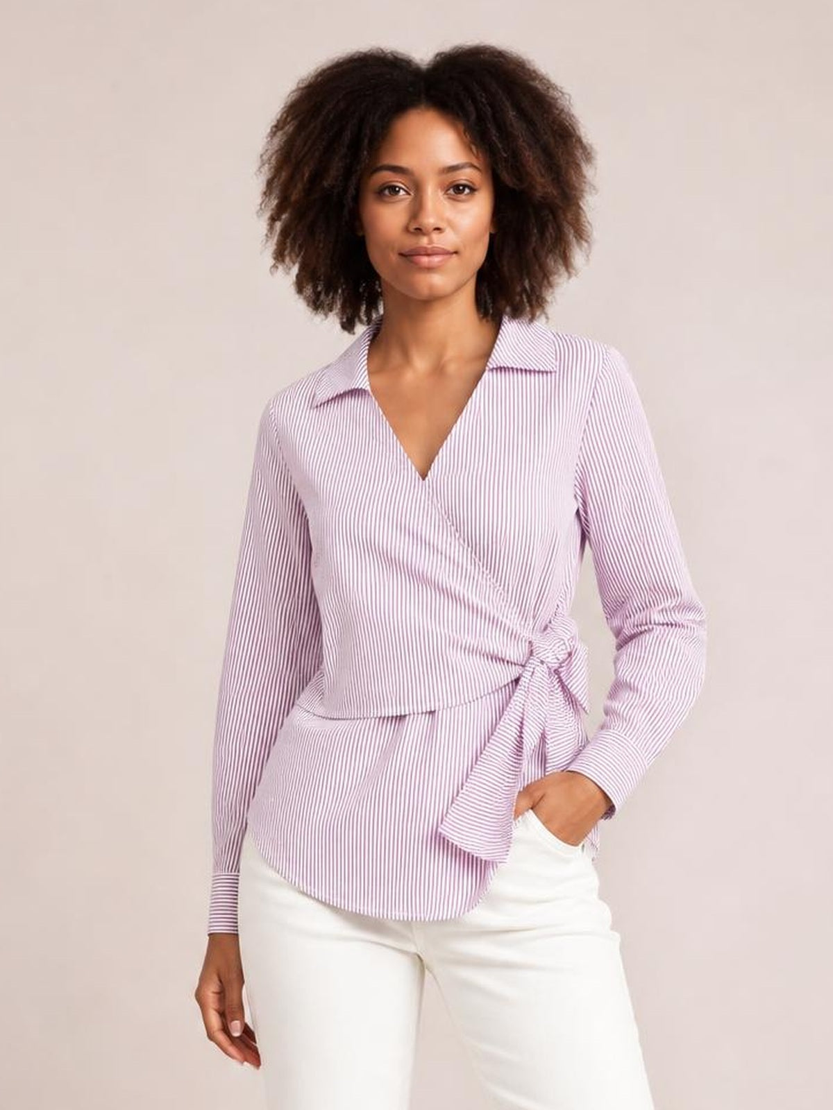 Model wearing a pink-and-white striped blouse with a Johnny collar, long sleeves, regular silhouette, and bow-tie waist detail, posing with one hand in her pocket.