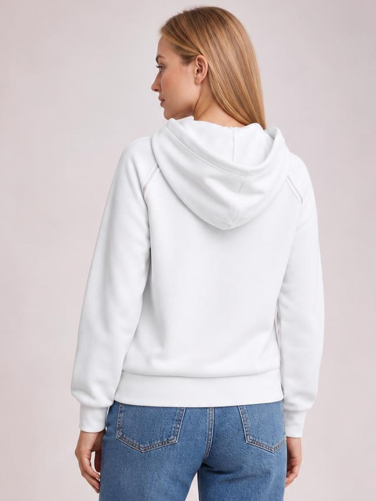 A woman models the white hoodie from the back, showing its relaxed silhouette, oversized hood, raglan sleeves, ribbed cuffs and hem, and contrast stitching.
