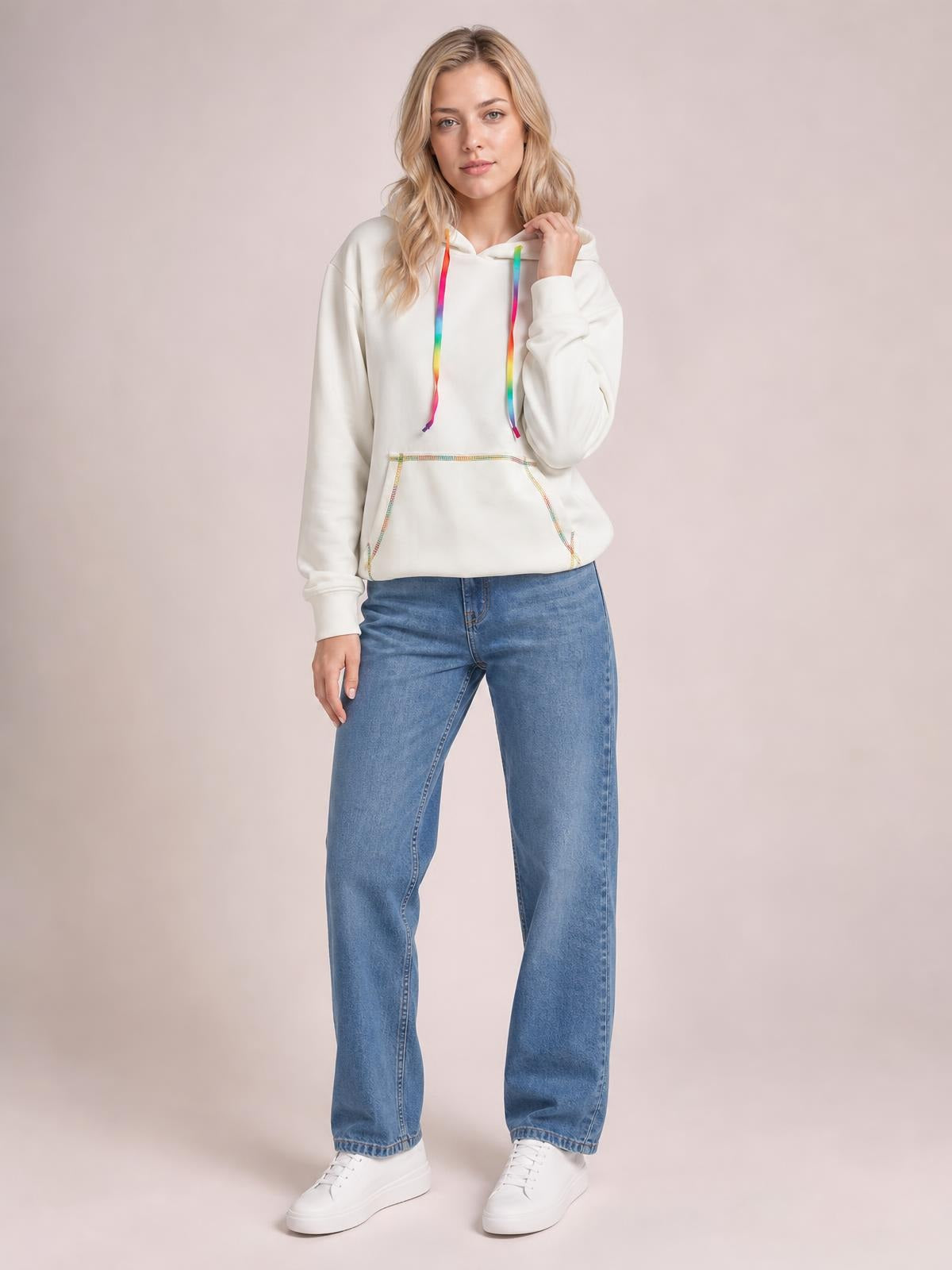 Model wearing an opaque white hoodie with contrast rainbow stitching, drawstring hood, raglan sleeves, and a relaxed silhouette, touching the hood.