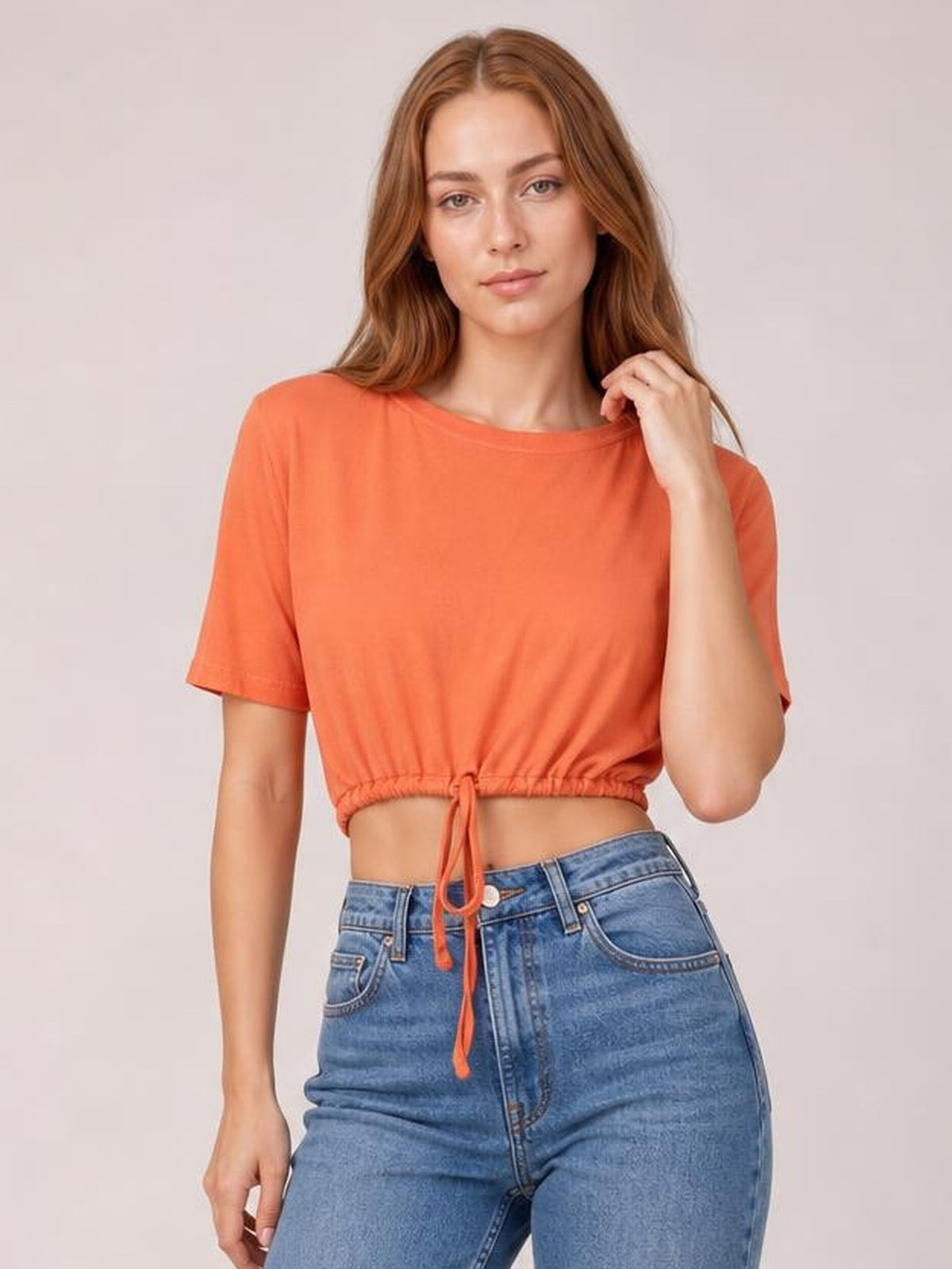 A model wears an orange drawstring round neck short sleeve T-shirt with a cropped silhouette, gathered hem, and relaxed fit while touching the neckline.