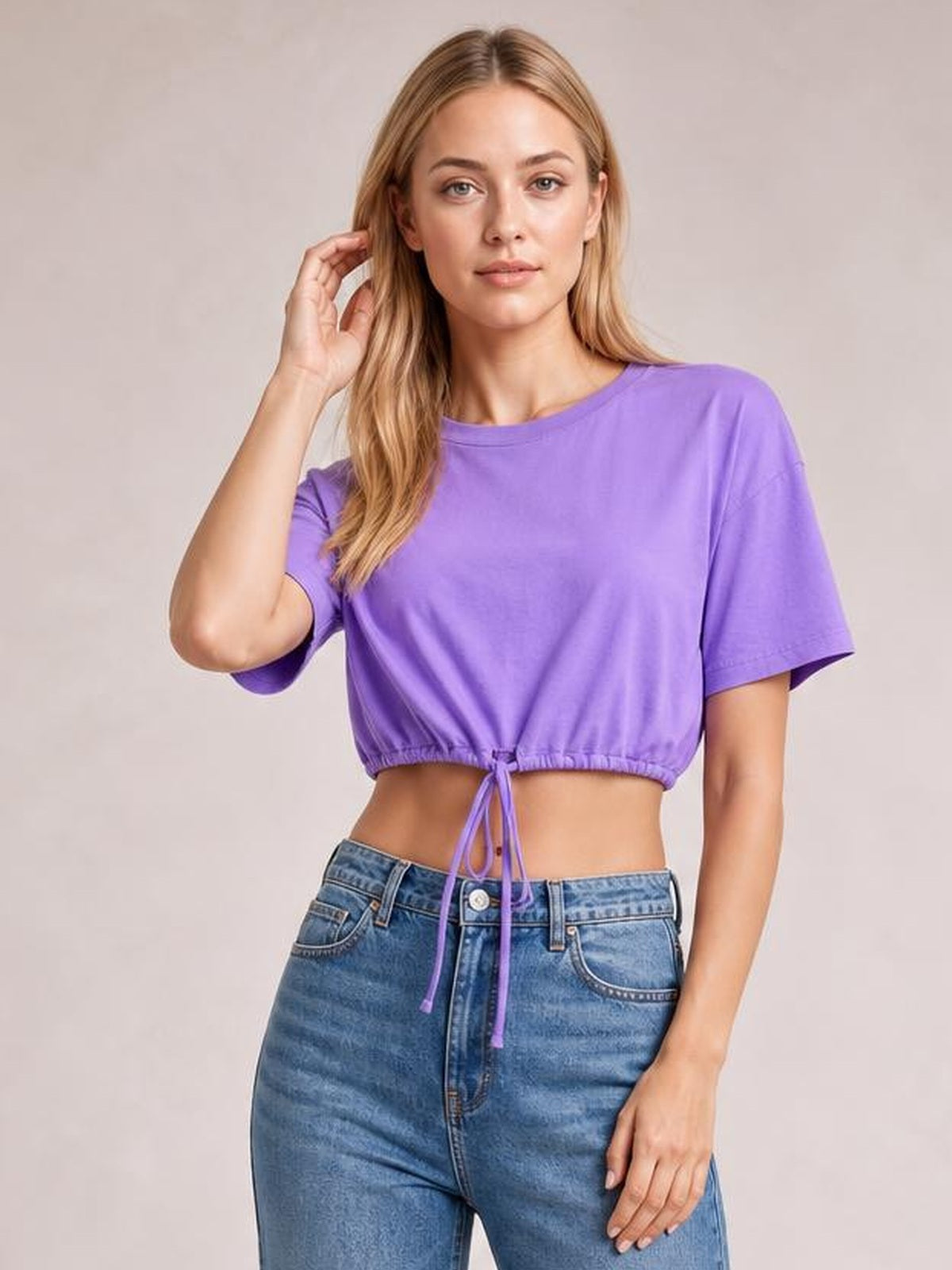 Model wearing a purple drawstring round neck short sleeve T-shirt with a cropped silhouette and adjustable tie hem, posing with one hand near her hair.