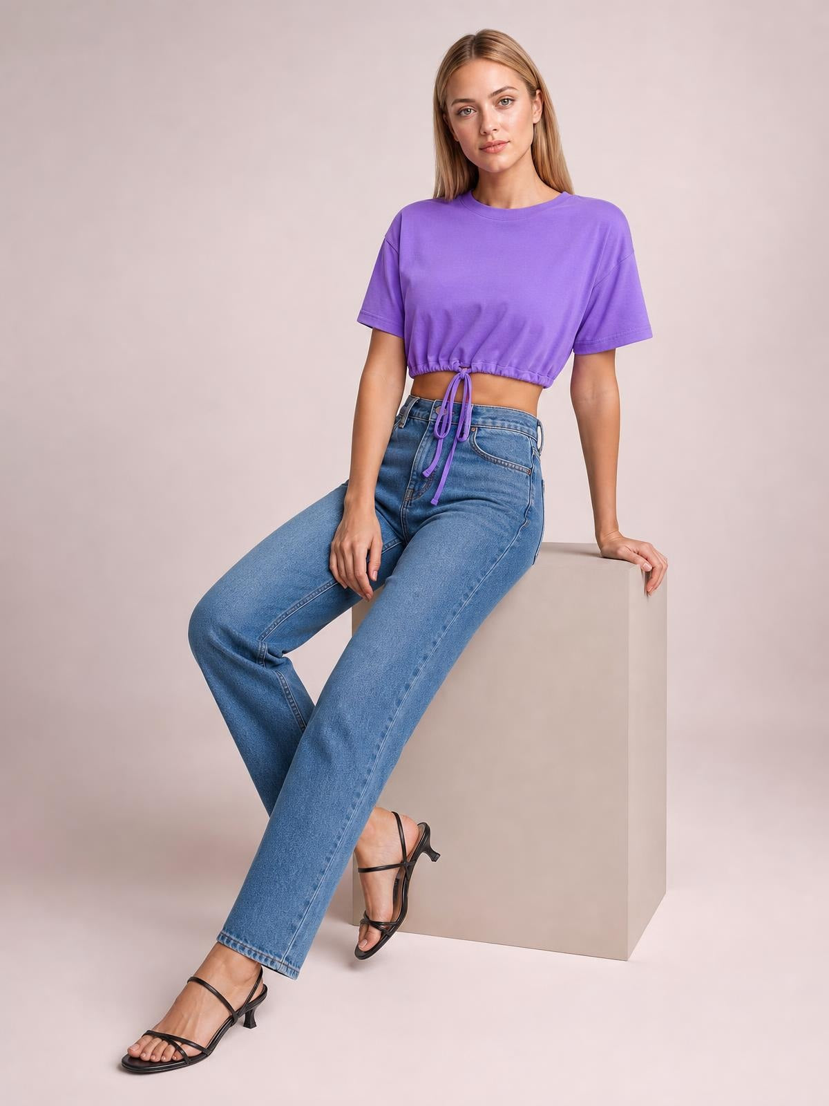 A model sits on a cube wearing a purple drawstring round-neck short-sleeve T-shirt with a cropped, adjustable hem and relaxed silhouette.