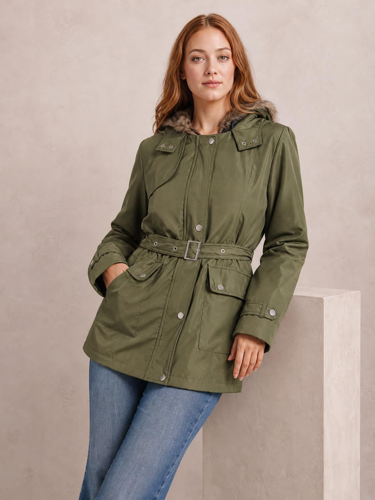 A woman poses in an olive hooded jacket with a faux-fur-trimmed collar, button front, belted waist, long sleeves, and multiple flap pockets.