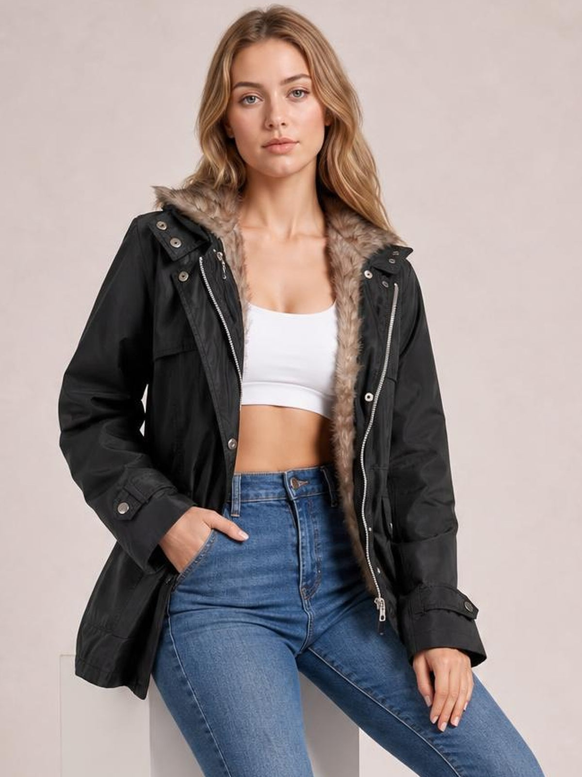 A model poses with one hand in her pocket wearing an open black hooded jacket with a plush detachable liner, zip and button closures, and a relaxed hip-length silhouette.