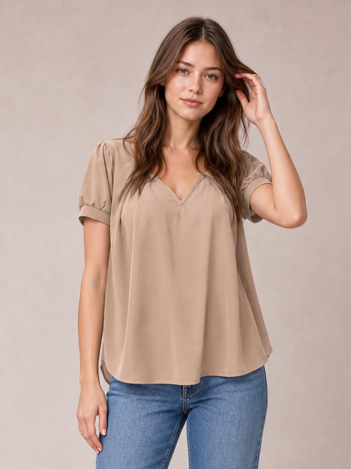 Model wearing a taupe notched short sleeve blouse with softly ruched shoulders, cuffed sleeves, and a relaxed, flowing silhouette while touching her hair.