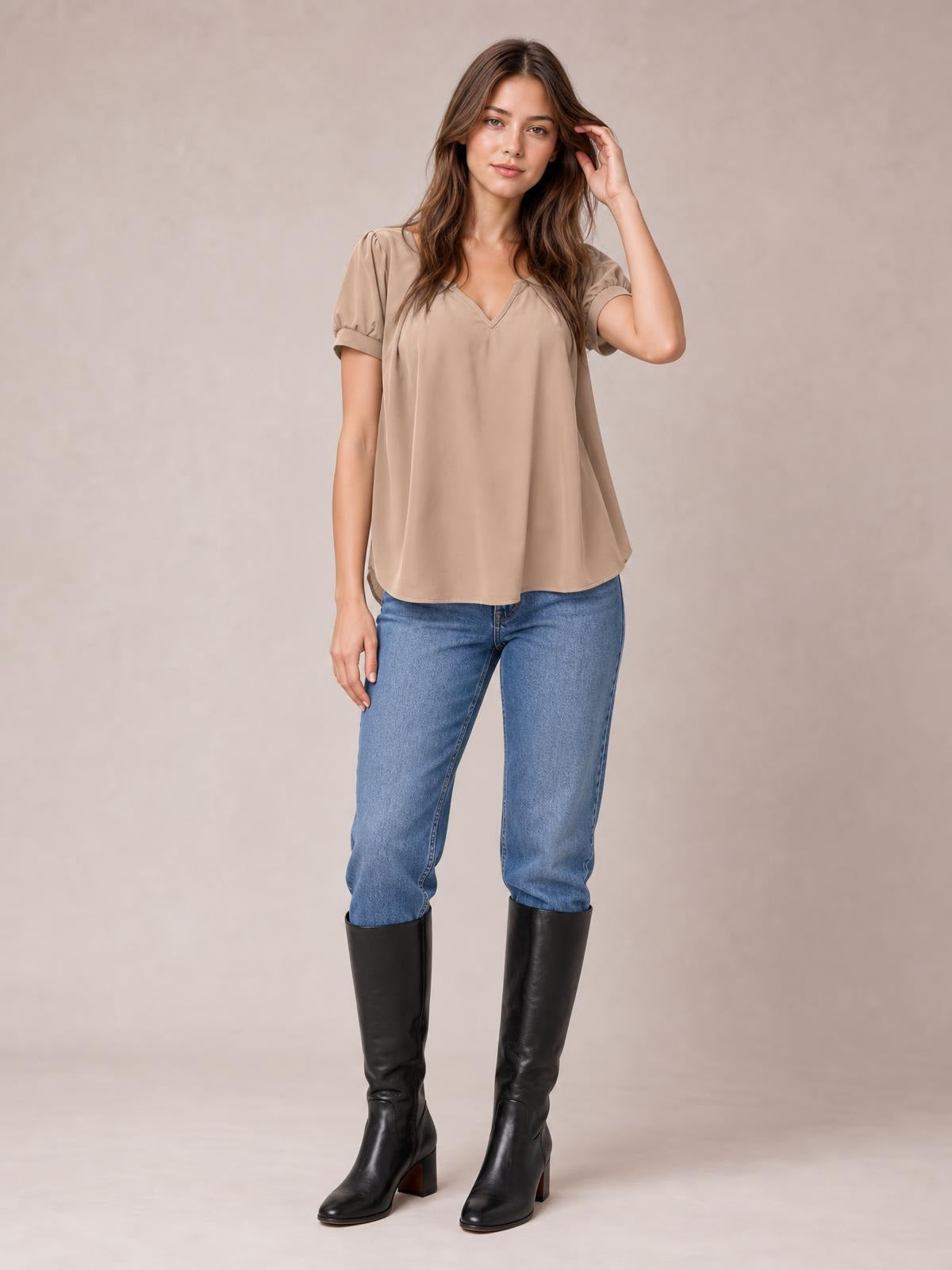 Model wearing a taupe notched short sleeve blouse with softly ruched shoulders, a relaxed flowy silhouette, and curved hem, posing with one hand in her hair.