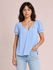 Model wears a light blue opaque blouse with a notched neckline, ruched shoulders, short puff sleeves, and a relaxed silhouette, posing with one hand in her pocket.