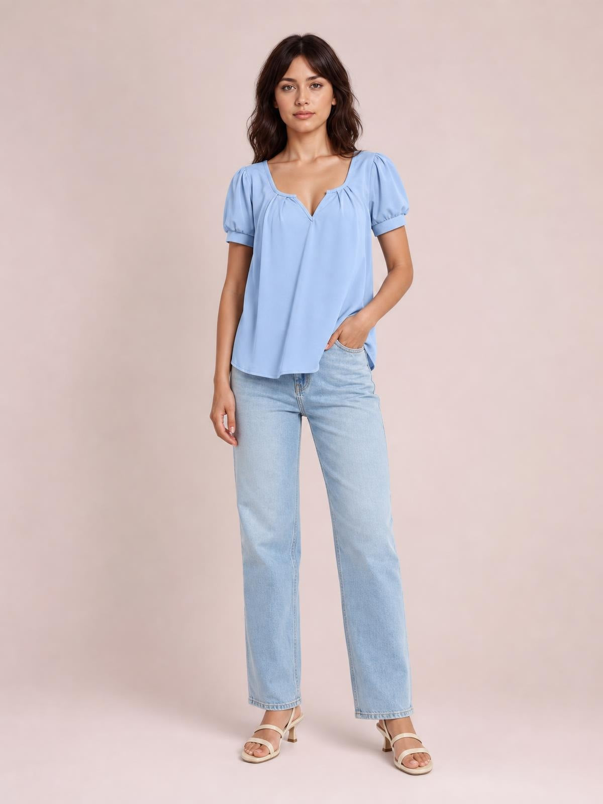 Model wears a light blue notched short-sleeve blouse with ruched shoulders, a relaxed flowing silhouette, and one hand resting in her jeans pocket.