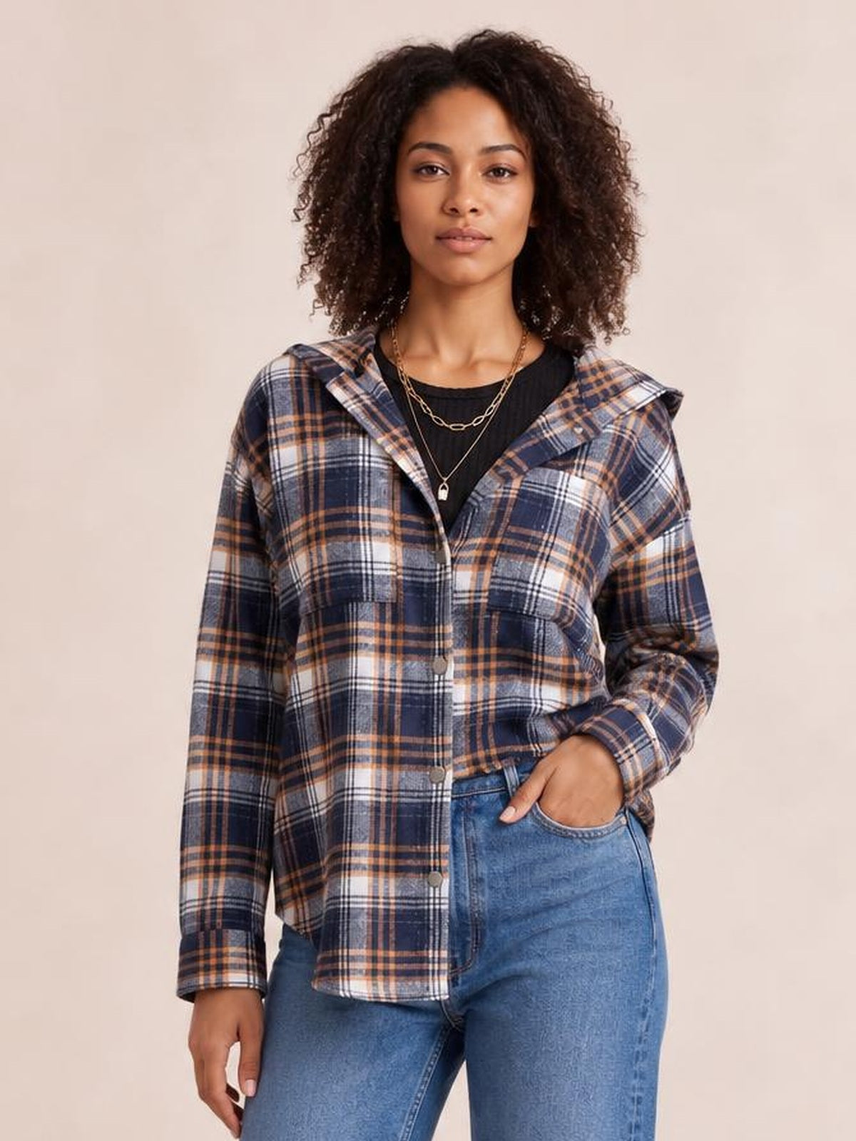 Model wears a navy, tan, and white plaid button-up hooded jacket with long sleeves, front pockets, and a relaxed silhouette, posing with one hand in a pocket.