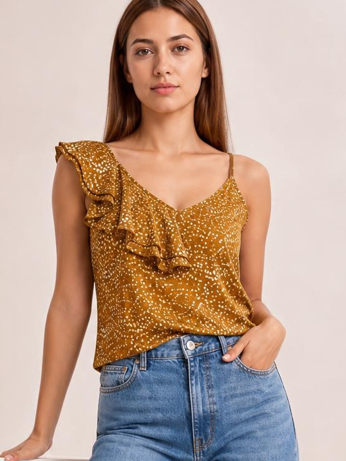 A model wears a mustard-gold polka dot asymmetrical tank top with spaghetti straps, a V-neckline, and layered ruffle trim across one shoulder.