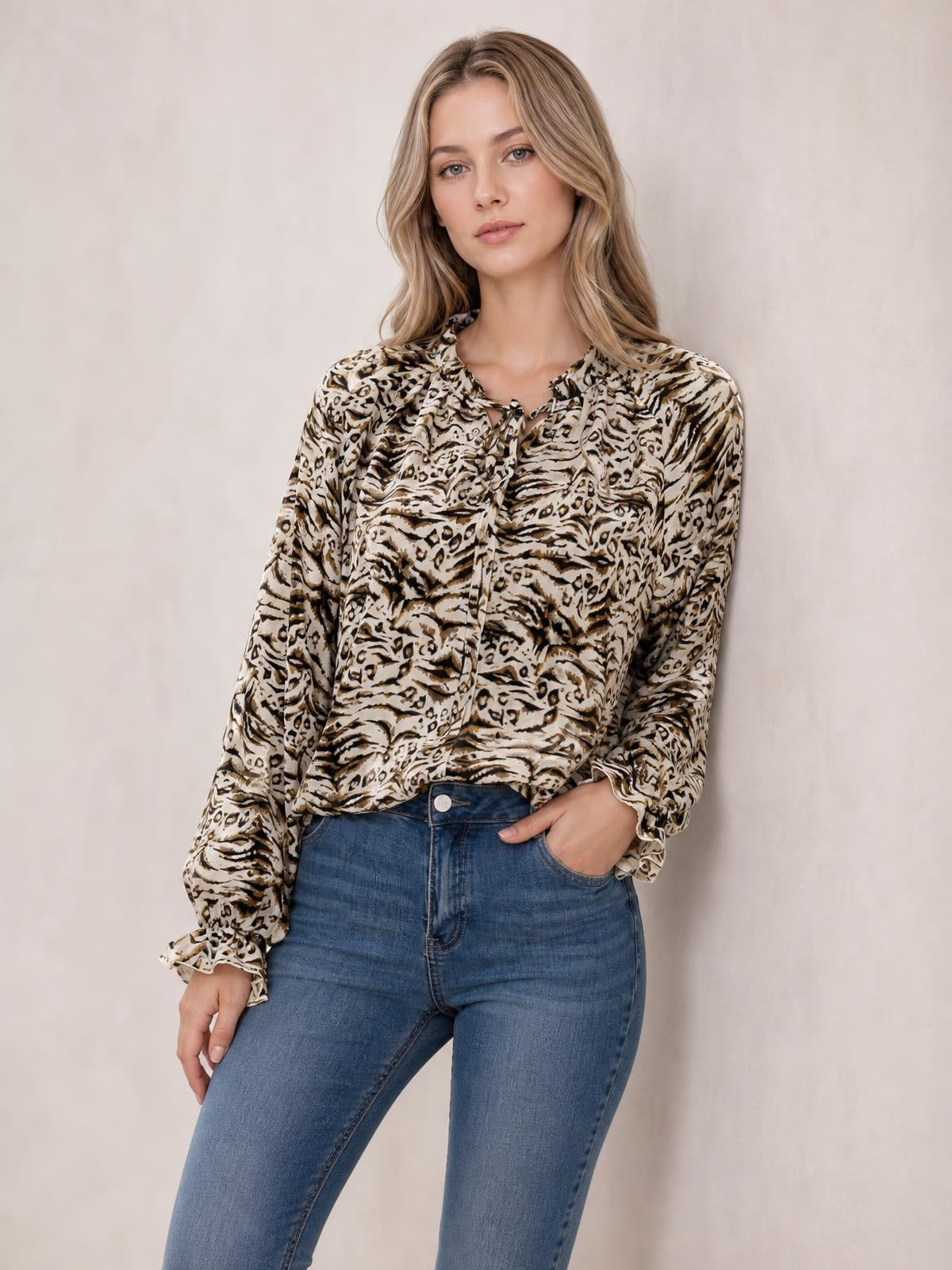 A woman poses with one hand in her jeans pocket, wearing a beige and black printed blouse with a tie neck, frill trim, and long flounce sleeves.