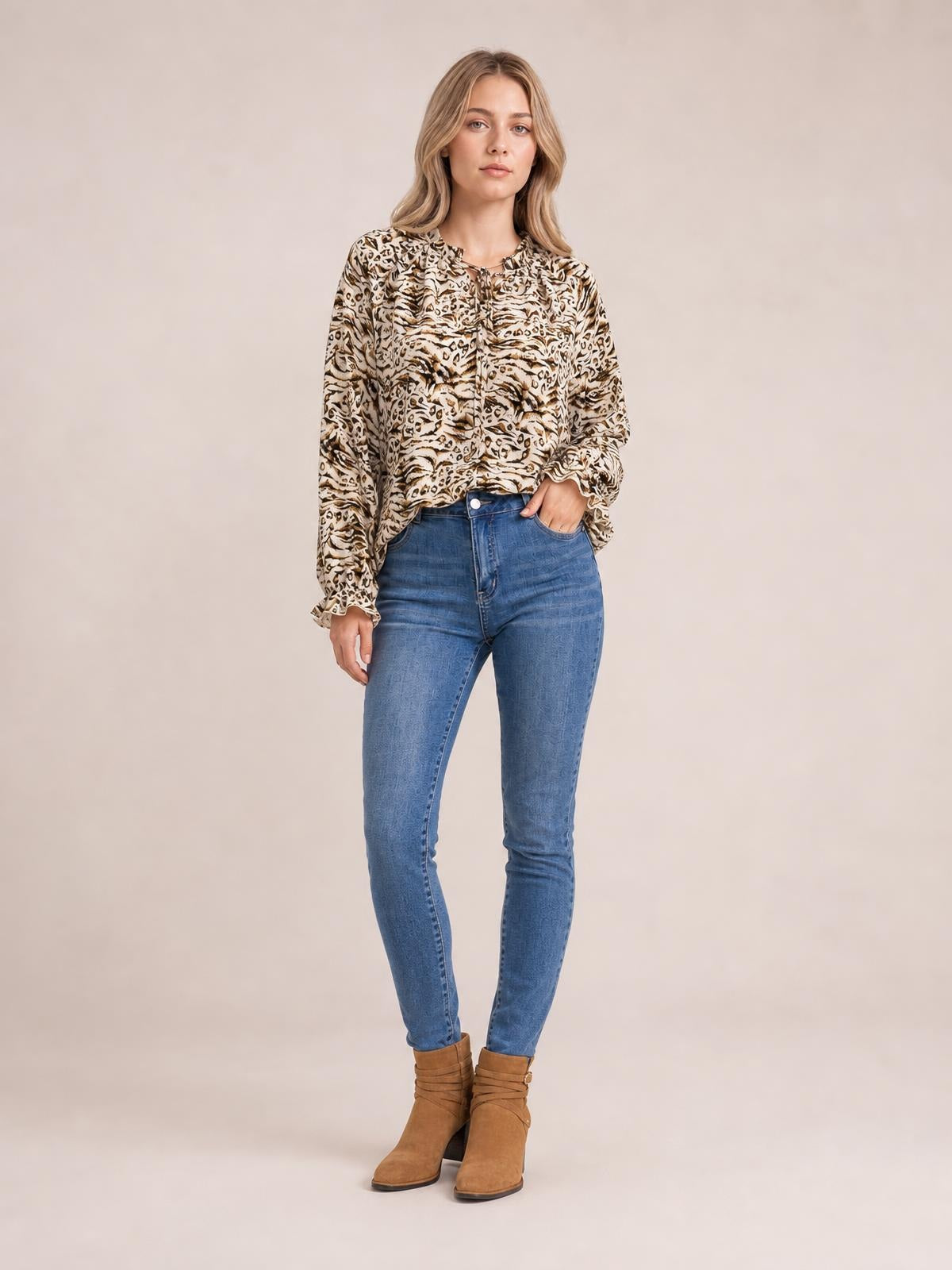 Model wearing a beige and brown printed tie-neck blouse with frill trim, long flounce sleeves, and a relaxed silhouette, standing with one hand in her pocket.