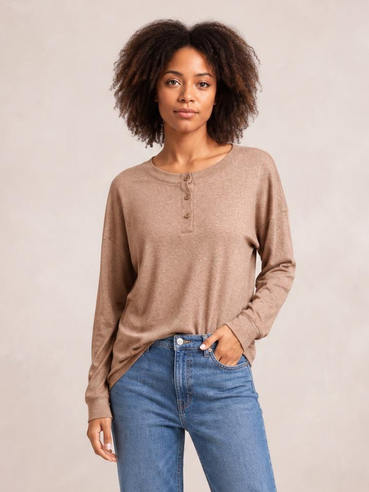 Model wearing a taupe quarter-button blouse with a crew neckline, dropped shoulders, long sleeves, and a relaxed silhouette, posing with one hand in her jeans pocket.