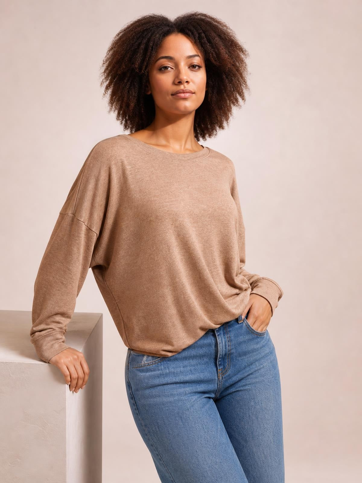 Model wears a taupe dropped-shoulder blouse with a round neckline, long sleeves, and a relaxed silhouette while leaning casually with one hand in her jeans pocket.