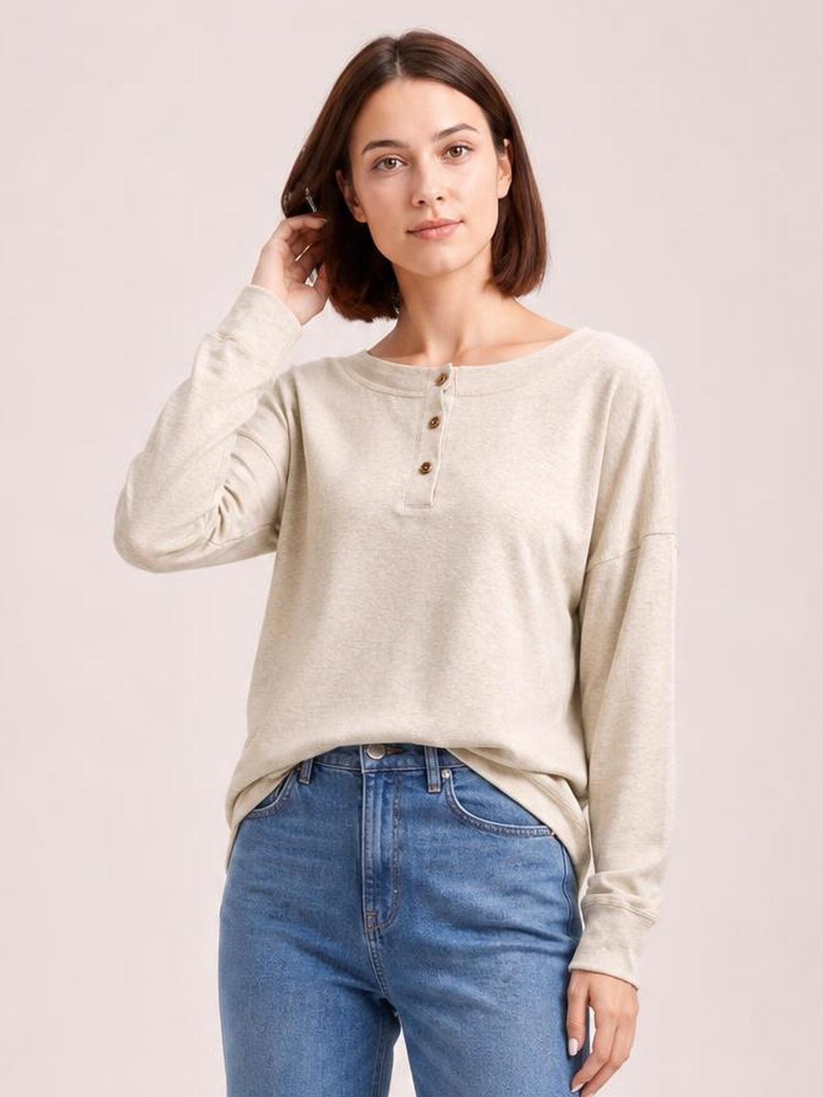 A model wears a light beige quarter-button dropped-shoulder blouse with long sleeves, a relaxed silhouette, and a round neckline while touching her hair.