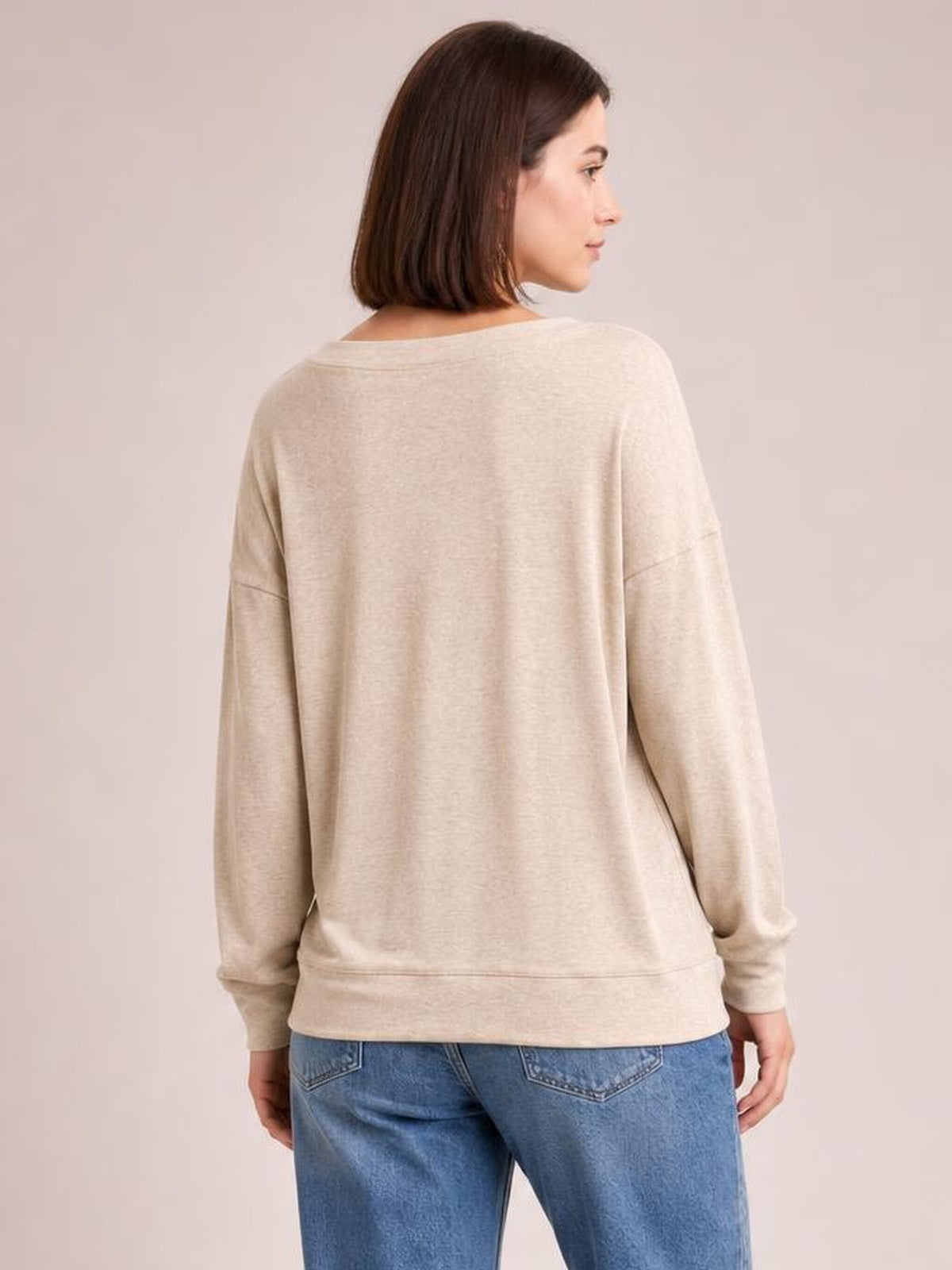 A model faces away wearing a light beige dropped shoulder blouse with a relaxed silhouette, long sleeves, ribbed cuffs, and a ribbed hem.