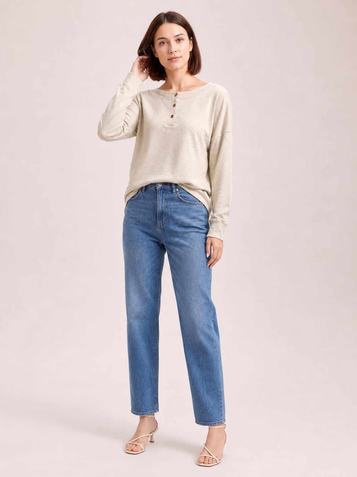 A woman wears a light beige quarter-button dropped-shoulder blouse with a round neckline, long sleeves, relaxed silhouette, and slightly curved hem.